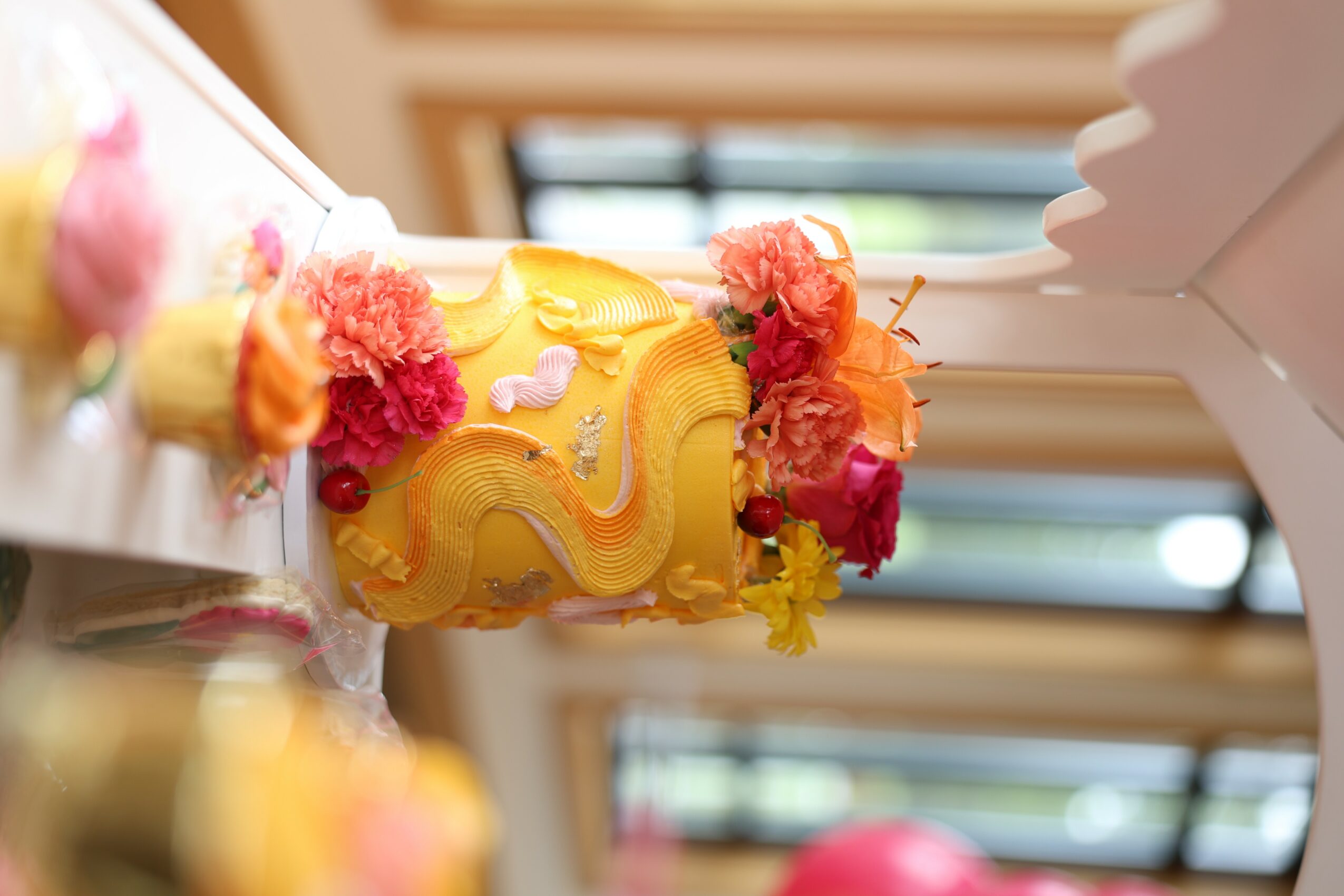 Decorated cake with yellow icing and colorful flowers, viewed from the side, with a window in the background.