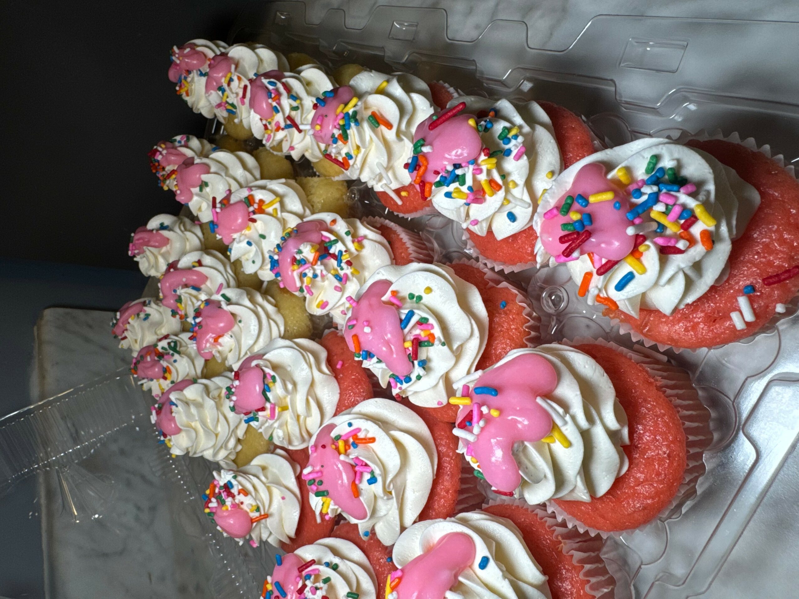 Red cupcakes with white frosting, pink decorations, and colorful sprinkles, arranged on a clear plastic tray.