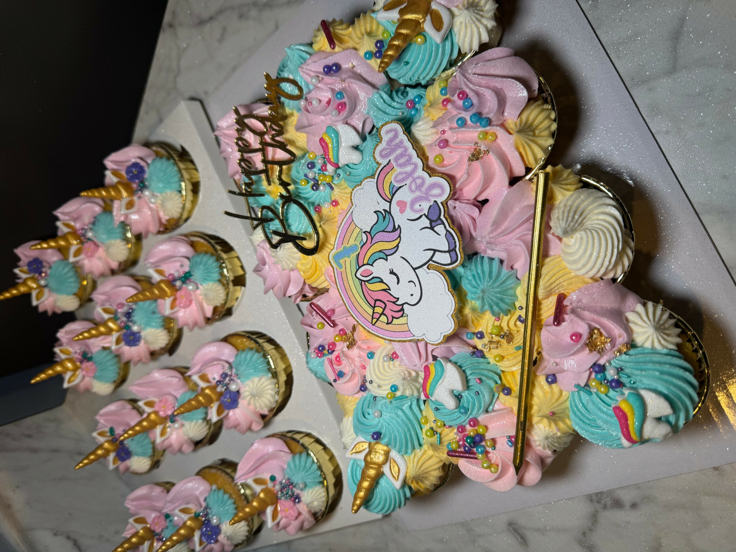 Unicorn-themed cake with pink, blue, and white frosting, decorated with unicorn figures, sprinkles, and gold accents, surrounded by cupcakes.