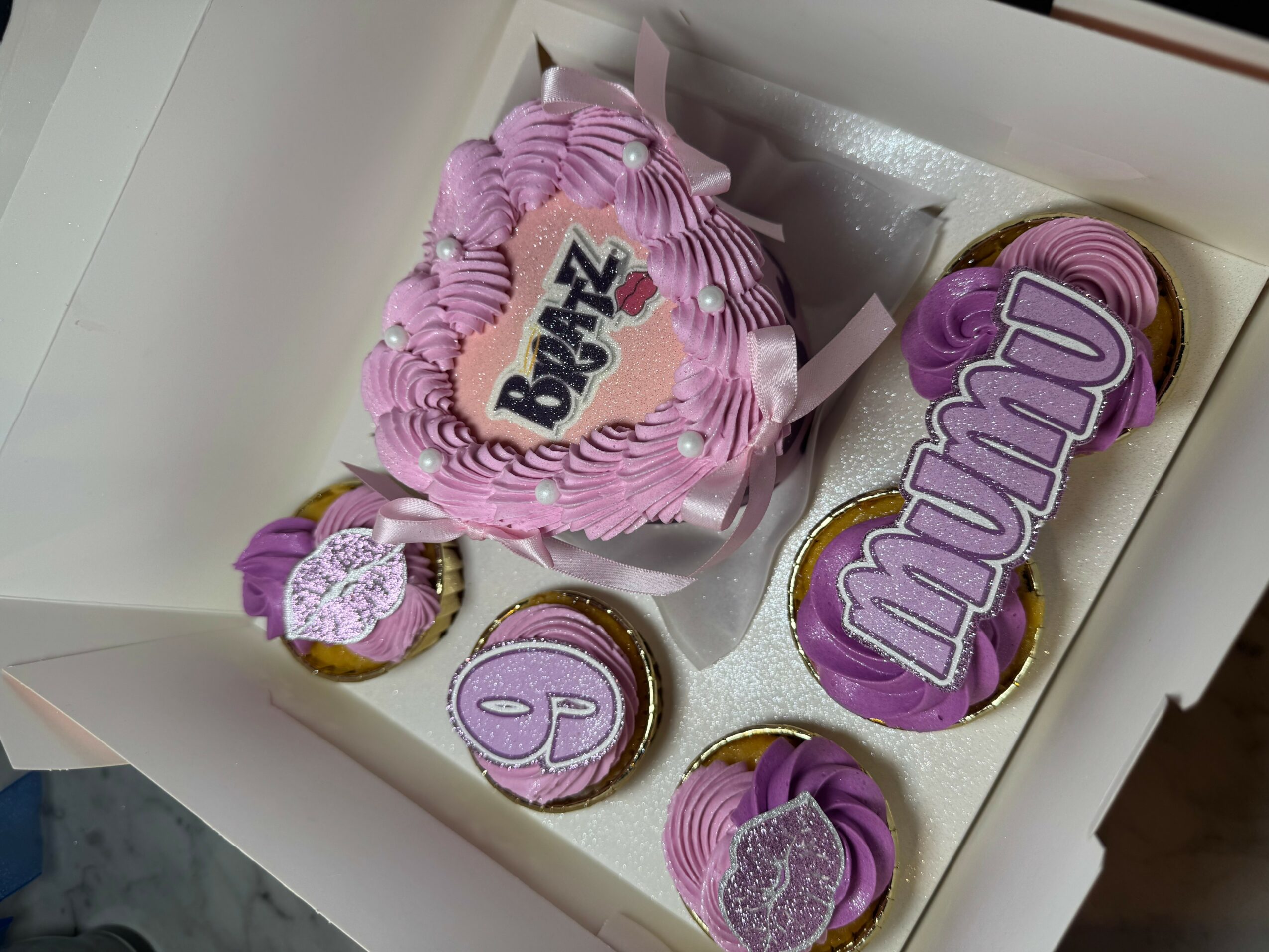 Box containing decorated cookies with purple and pink icing, some with heart shapes and text, on a white surface.