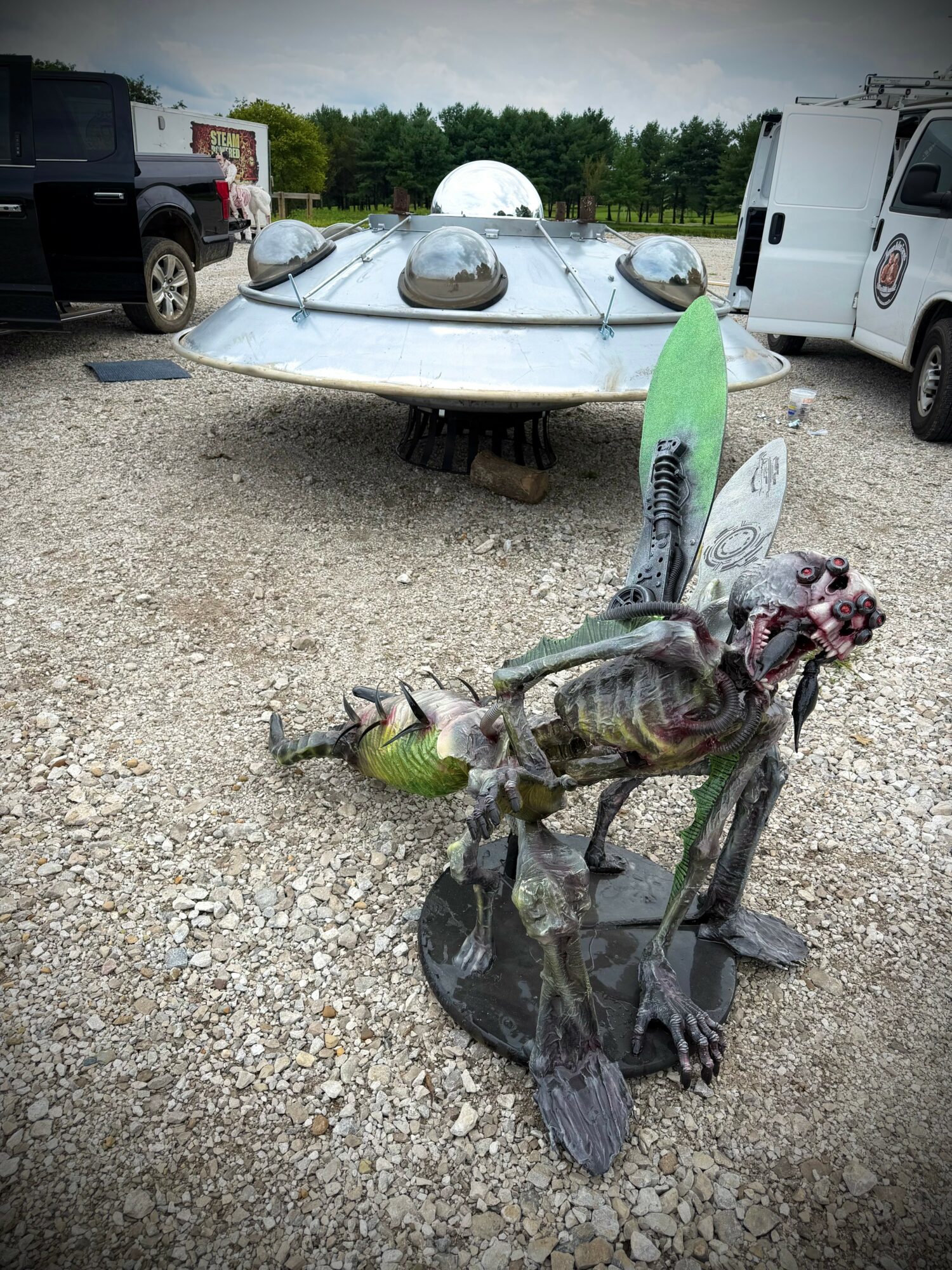 Alien spaceship with three domes on a gravel lot, surrounded by vehicles, and a dragon sculpture in the foreground.