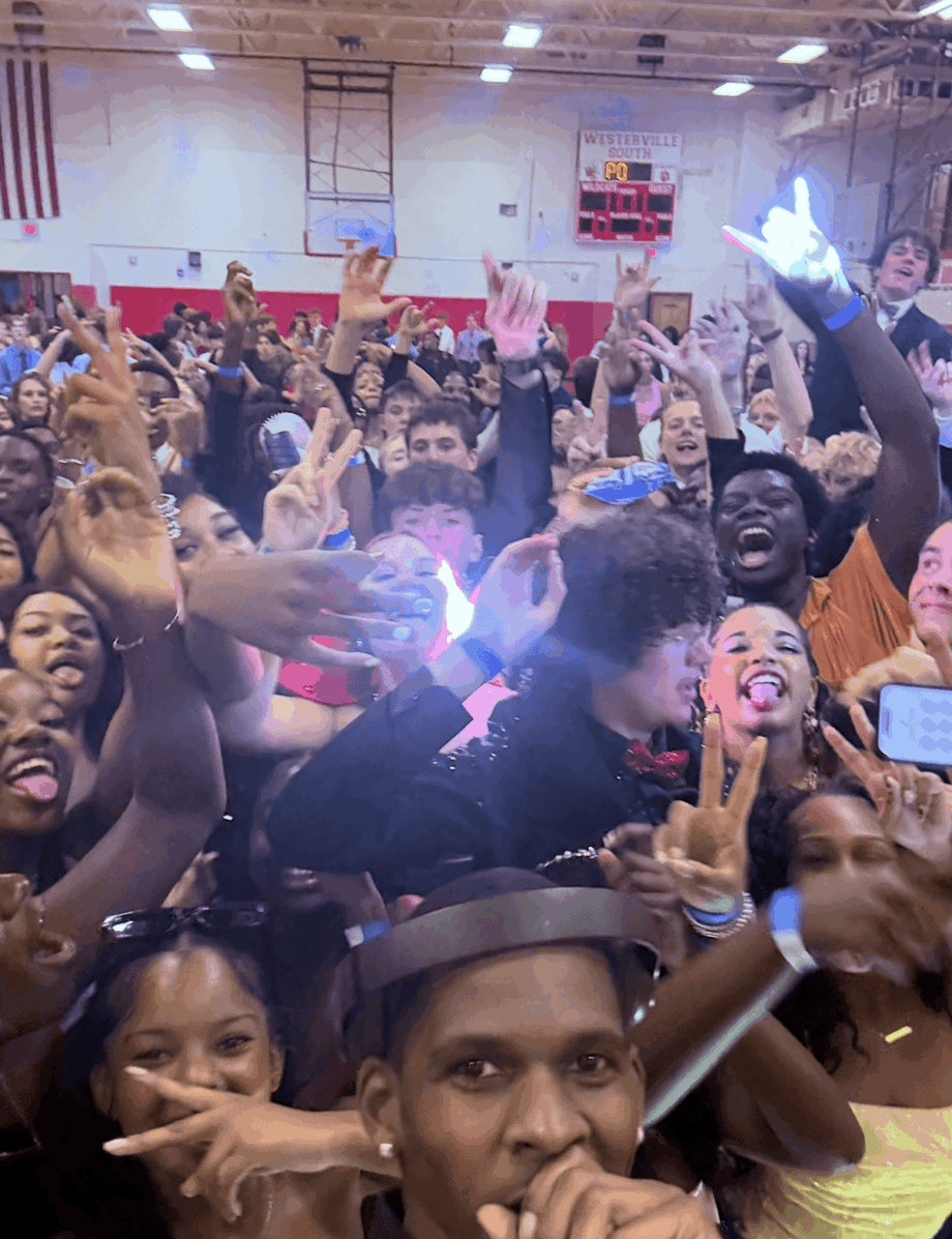 Crowd of people in a gymnasium, many smiling and raising hands, with some holding phones, bright lighting, American flag, and banners.