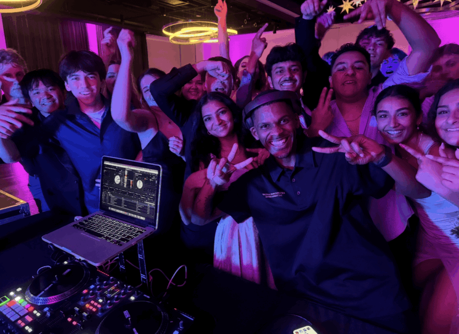 Group of people at a party or event, smiling and raising their hands, with a DJ setup in the foreground.