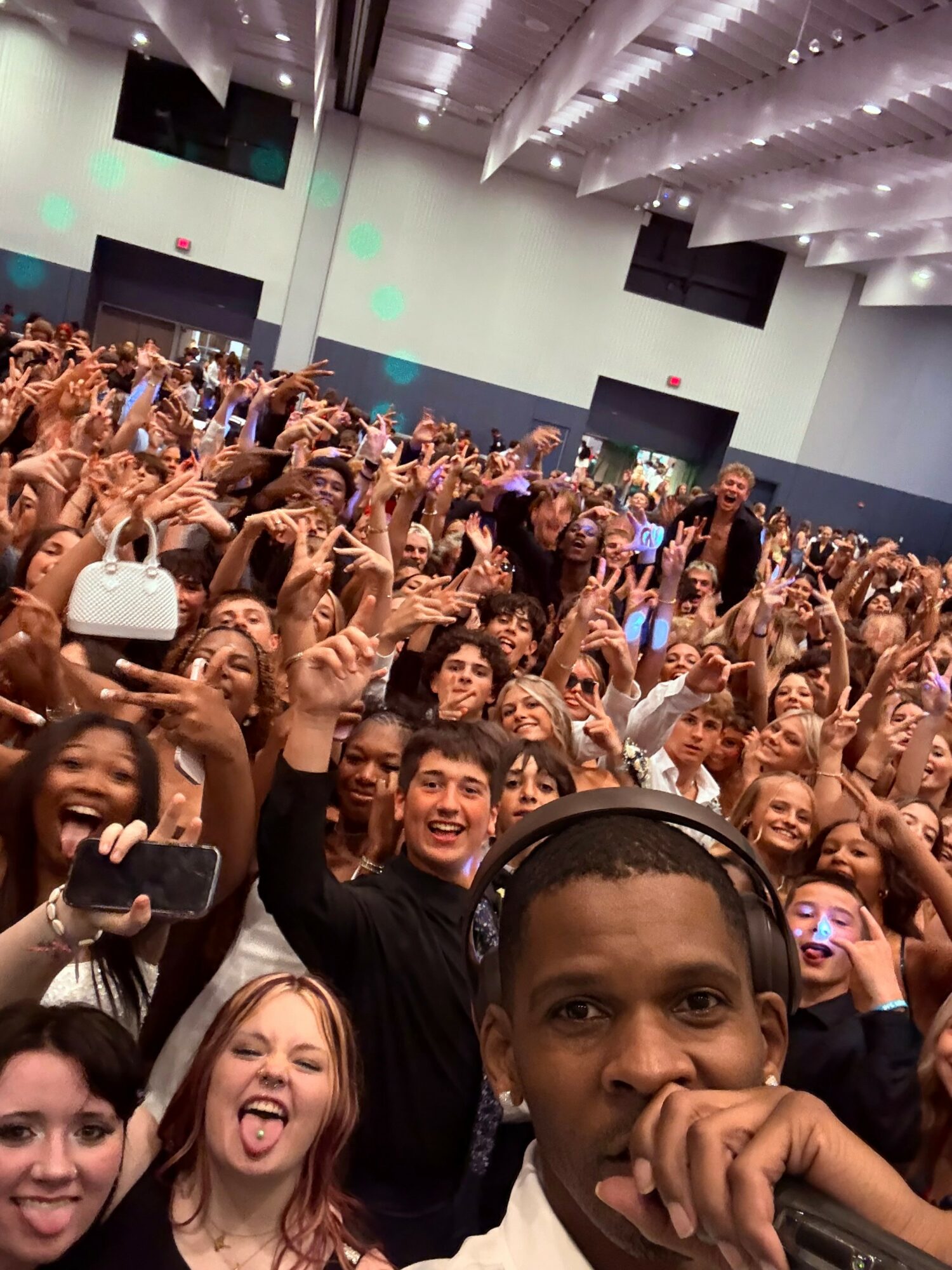 Crowd of people at a large indoor event, many smiling and raising hands, with a person taking a selfie in the foreground.