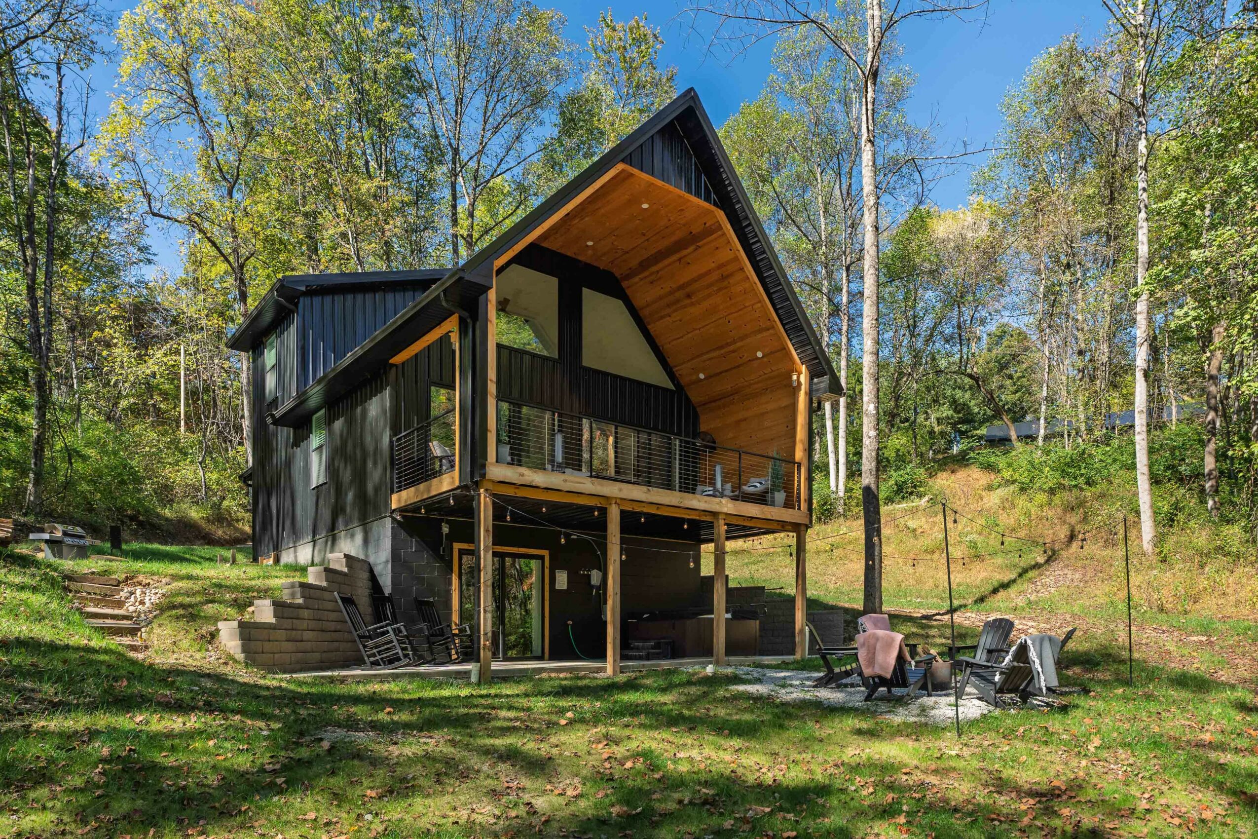 Modern house with a curved roof, large windows, and a balcony, surrounded by trees and outdoor seating.