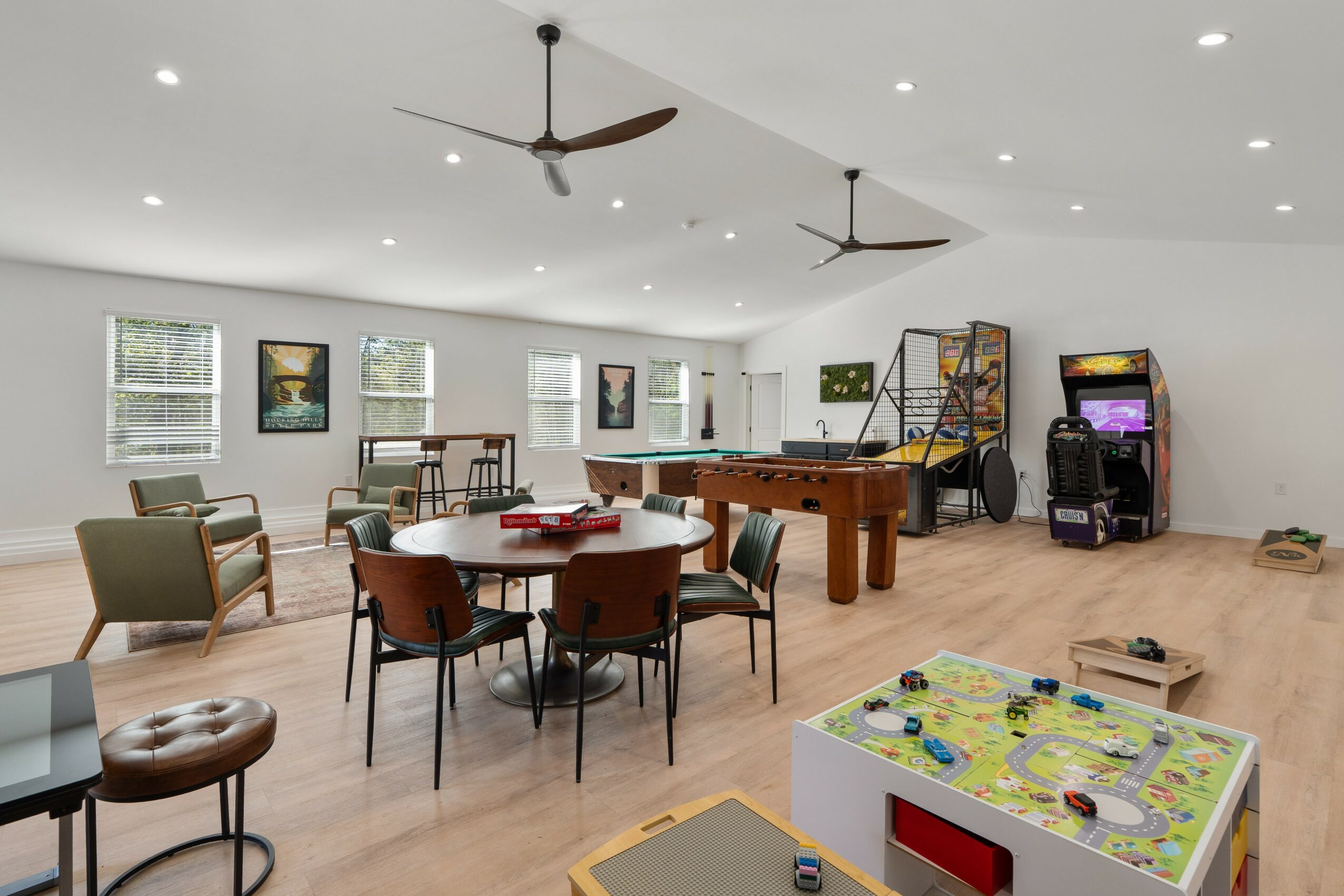 Game room with pool table, arcade machines, round table with chairs, and a colorful toy area, bright with windows and ceiling fans.