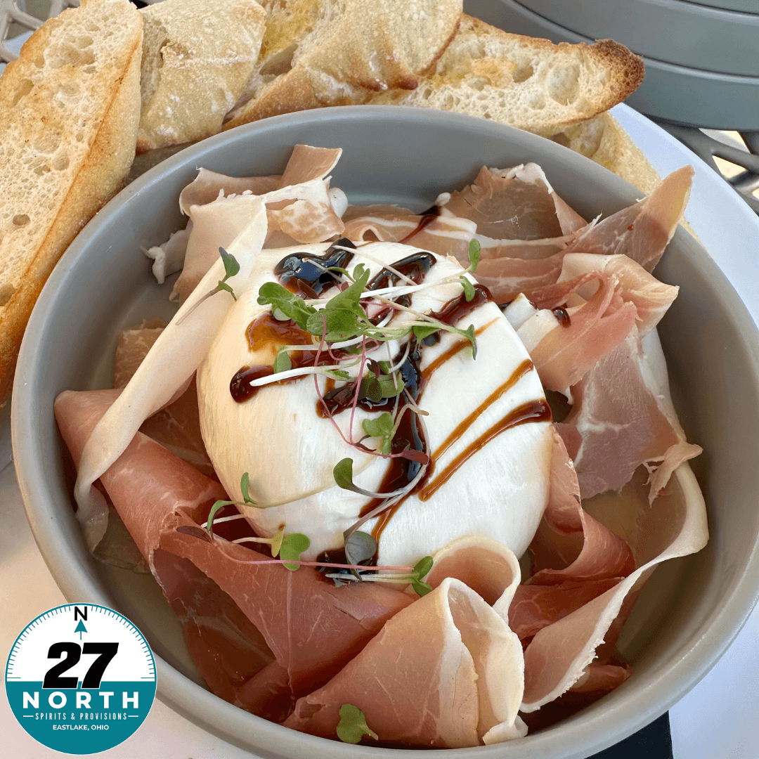 Bowl with prosciutto, cheese, a soft-boiled egg, microgreens, balsamic glaze, and bread slices on the side.