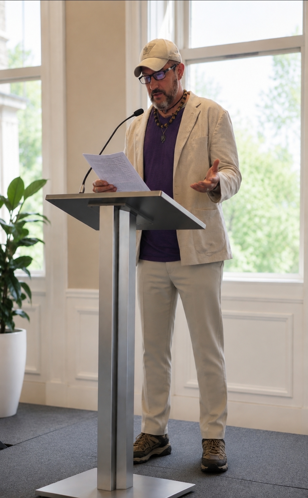 Man in beige suit, purple shirt, and cap speaking at a podium with large window behind him.