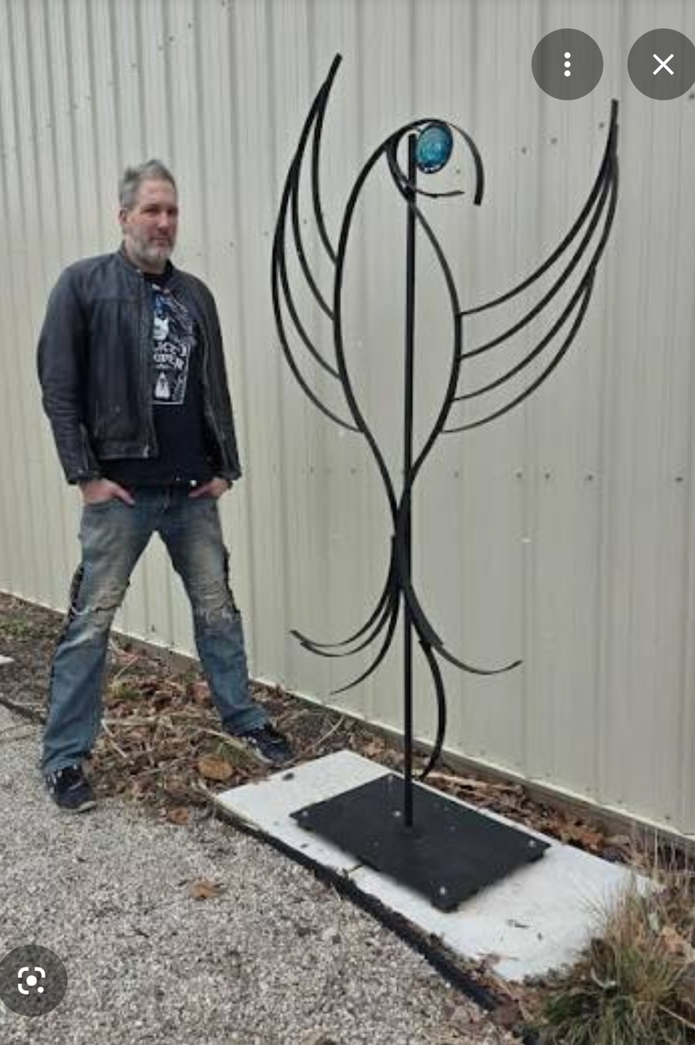 Man standing next to a tall metal sculpture resembling a stylized bird with wings, outdoors near a metal wall.
