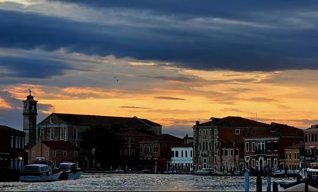 Sunset over a cityscape with buildings and a body of water, dark clouds in the sky, and a lighthouse in the distance.