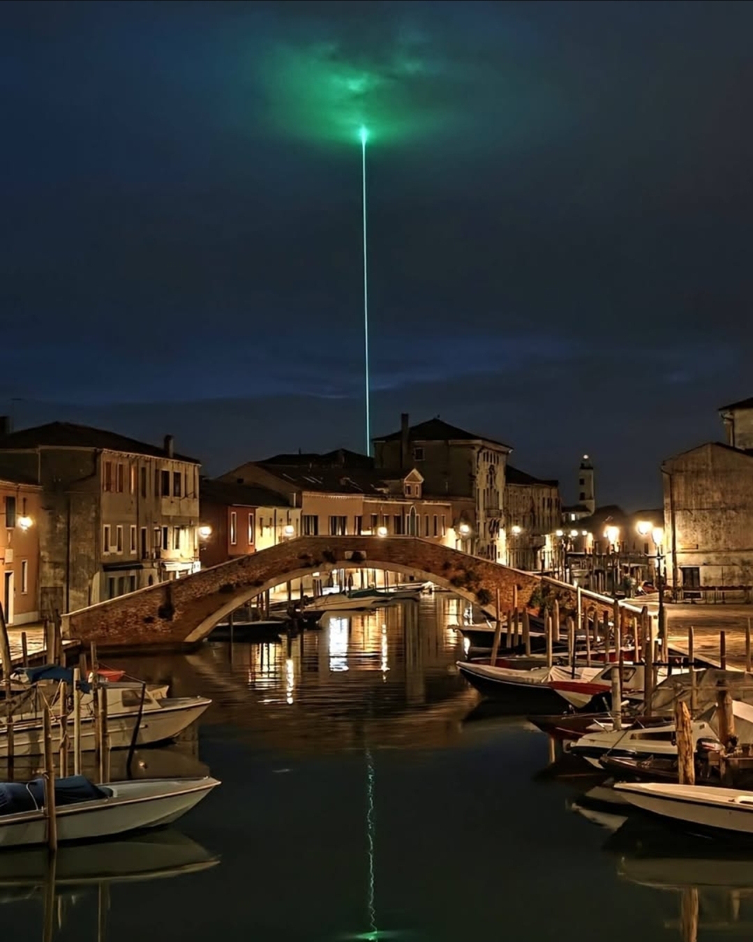 Night scene with boats docked in a canal, a bridge, buildings, and a green laser beam in the sky reflecting on the water.