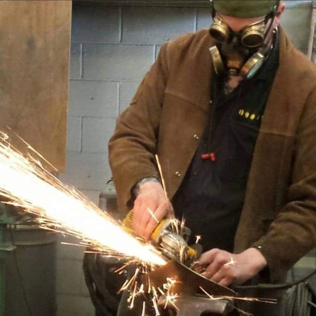 Person wearing goggles and a brown jacket using a grinder, sparks flying, in an industrial setting.