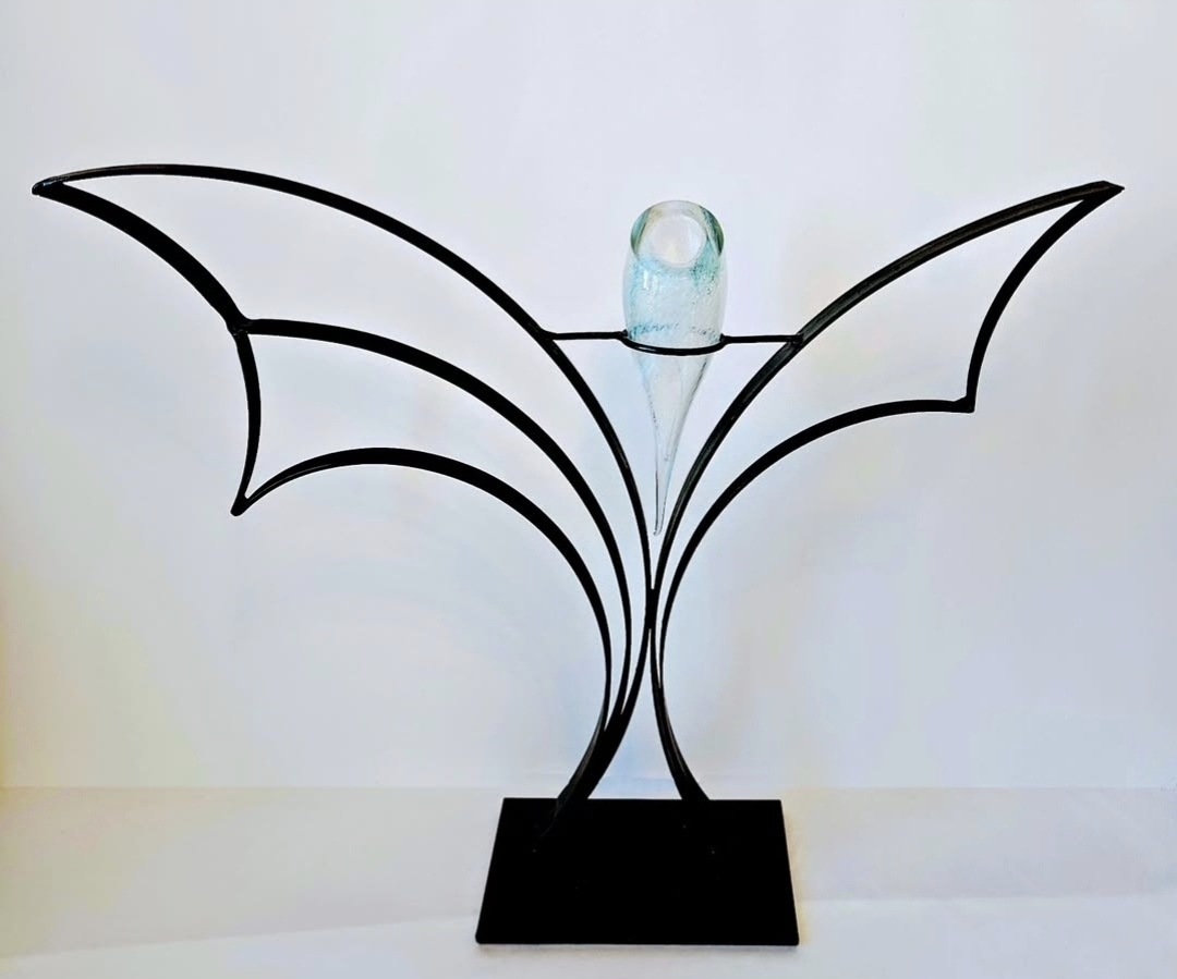 Metal structure resembling a bat with outstretched wings, holding a glass on top, on a black base.