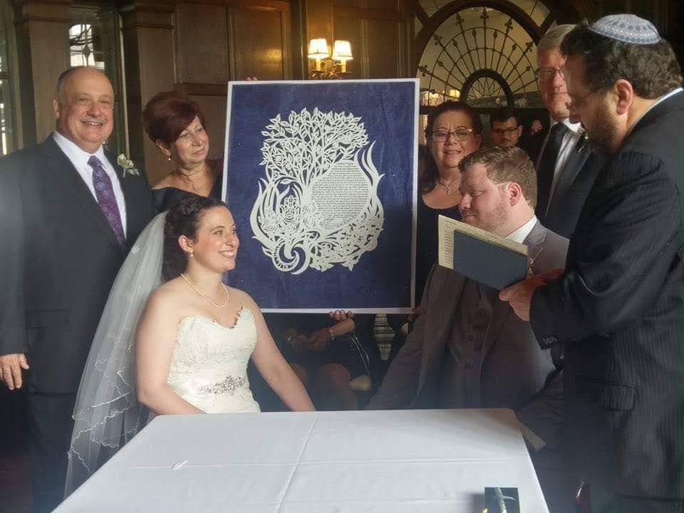 Group of people gathered around a bride and groom at a wedding ceremony, with a decorative artwork in the background.