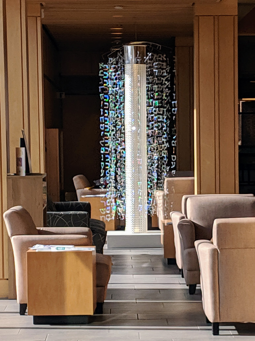 Interior space with chairs and a tall illuminated cylindrical display with floating letters and symbols, wooden walls, and a staircase.