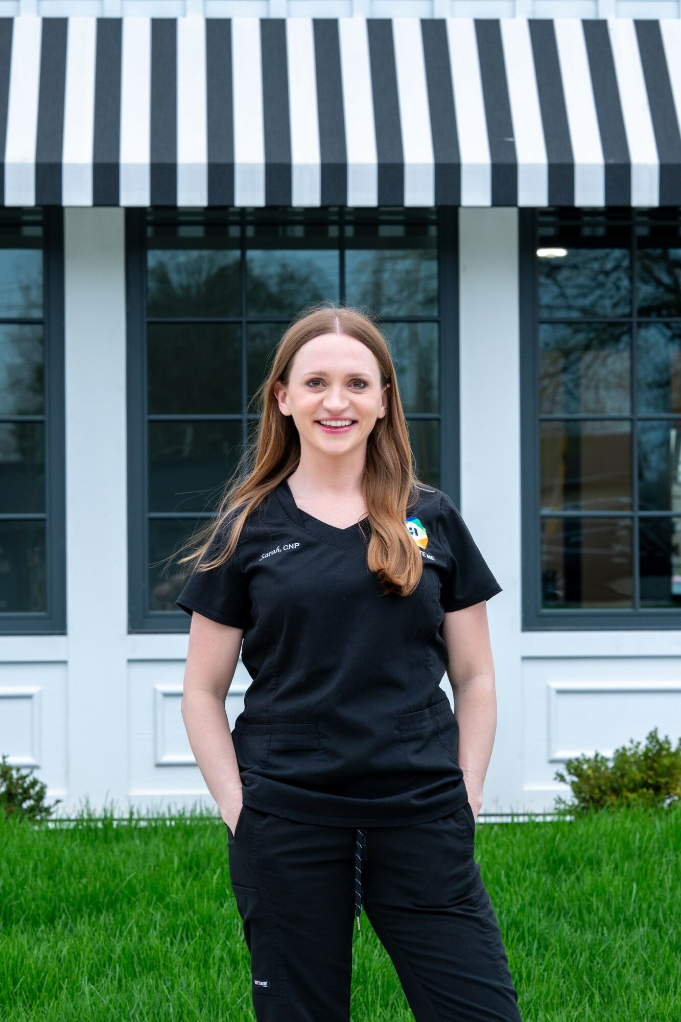 Young woman with long hair smiling outdoors in front of a building with large windows and a striped awning.