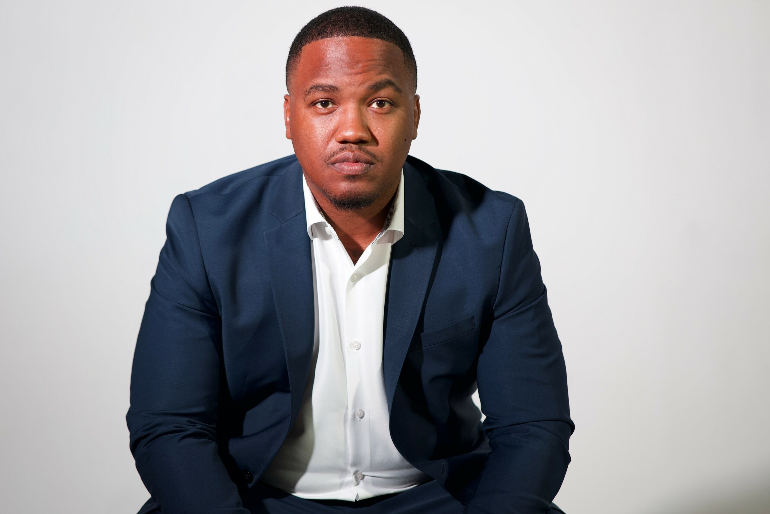 Man in a navy suit and white shirt sitting against a plain light background.
