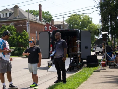 Three people standing outside near a black mobile unit on a grassy area, with houses and trees in the background.