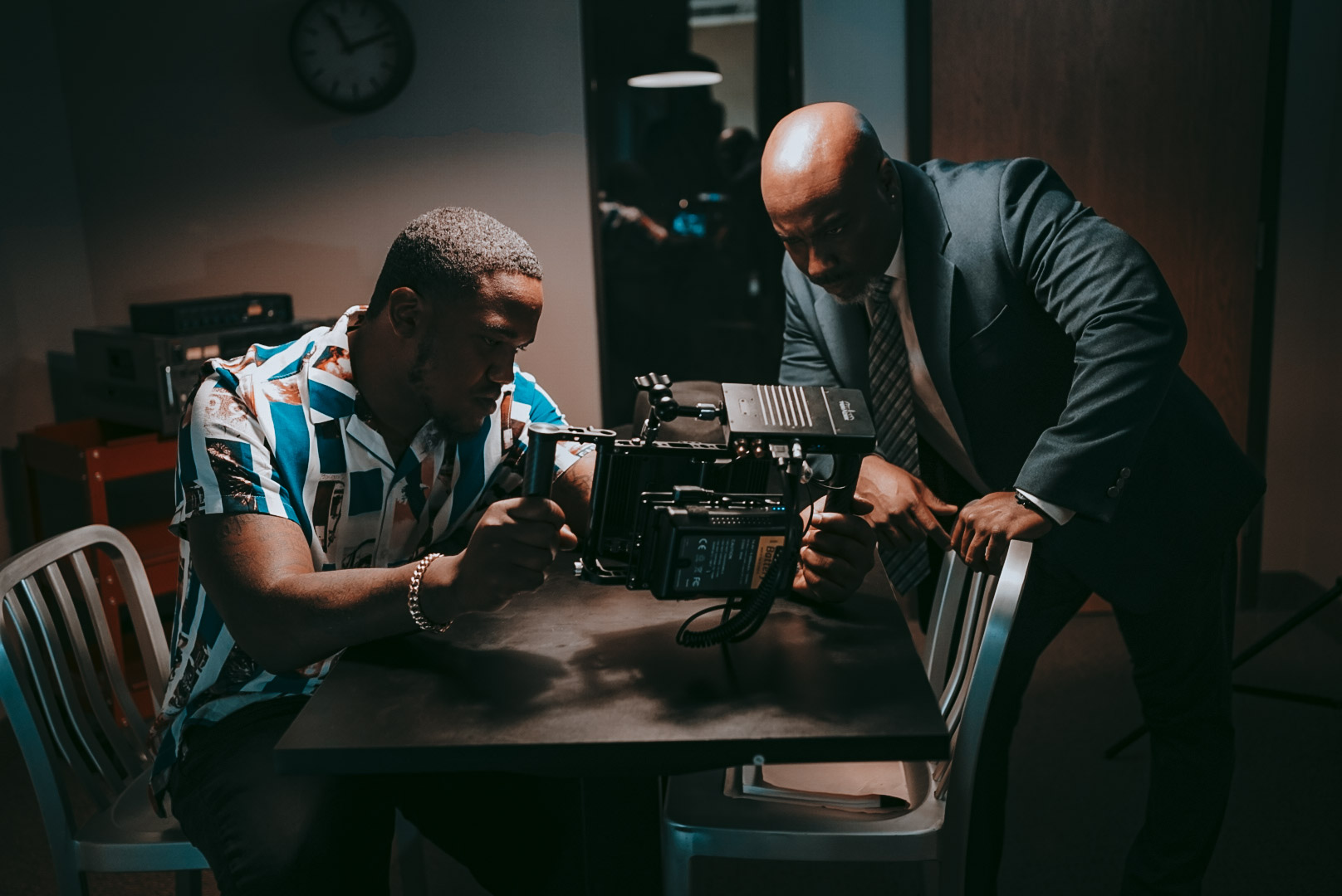 Two men examine a camera on a table in a dimly lit room, one seated and one standing, with a clock on the wall.