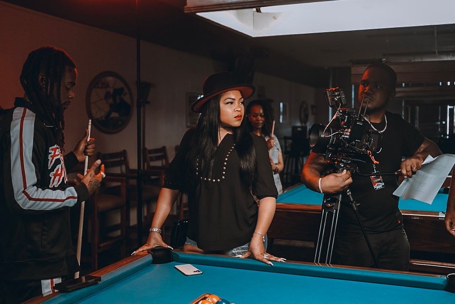 Group of people around a pool table in a dimly lit room, one woman wearing a hat, others watching.