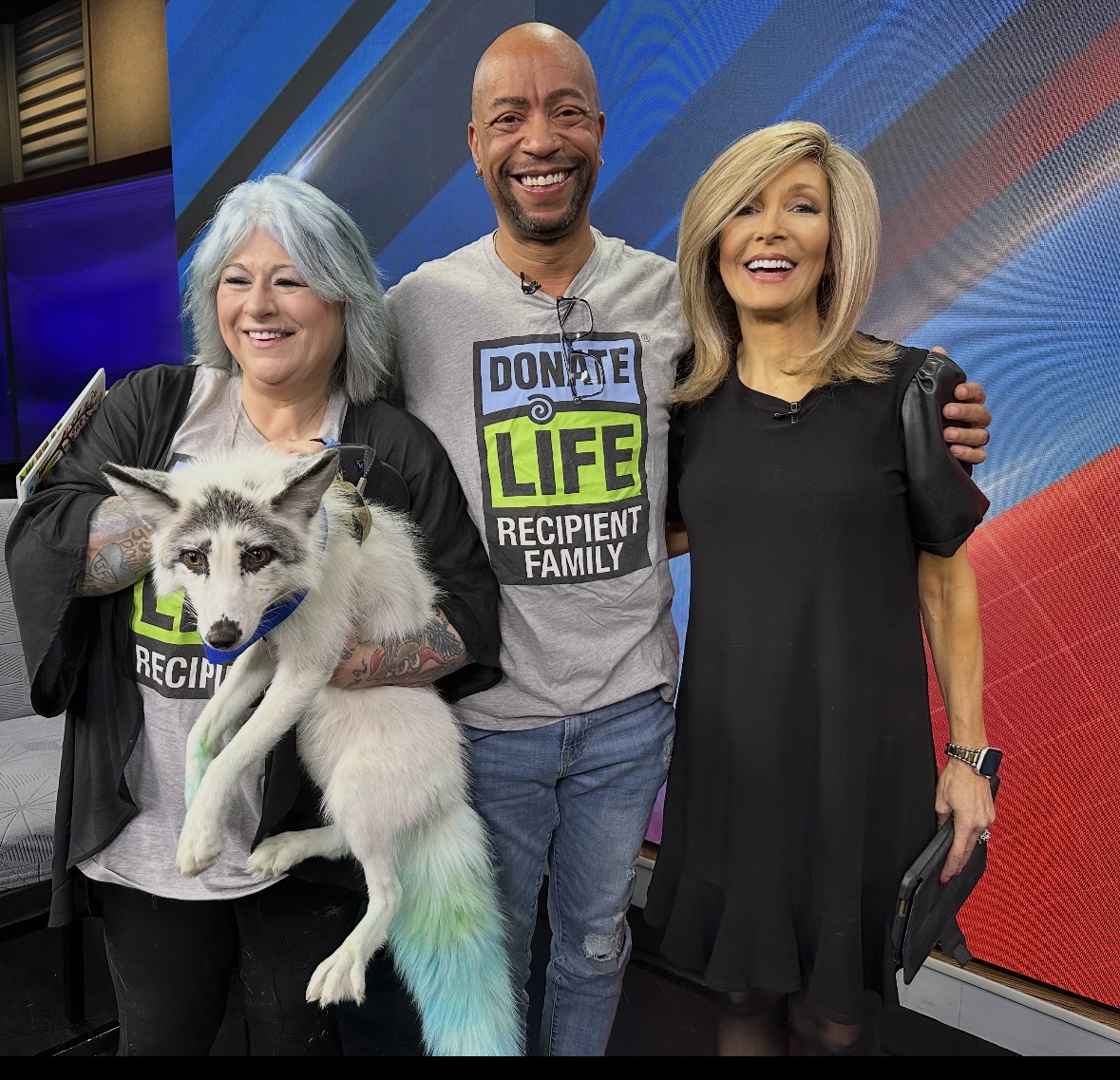 Three people smiling, one holding a puppy, in a studio setting with colorful background.