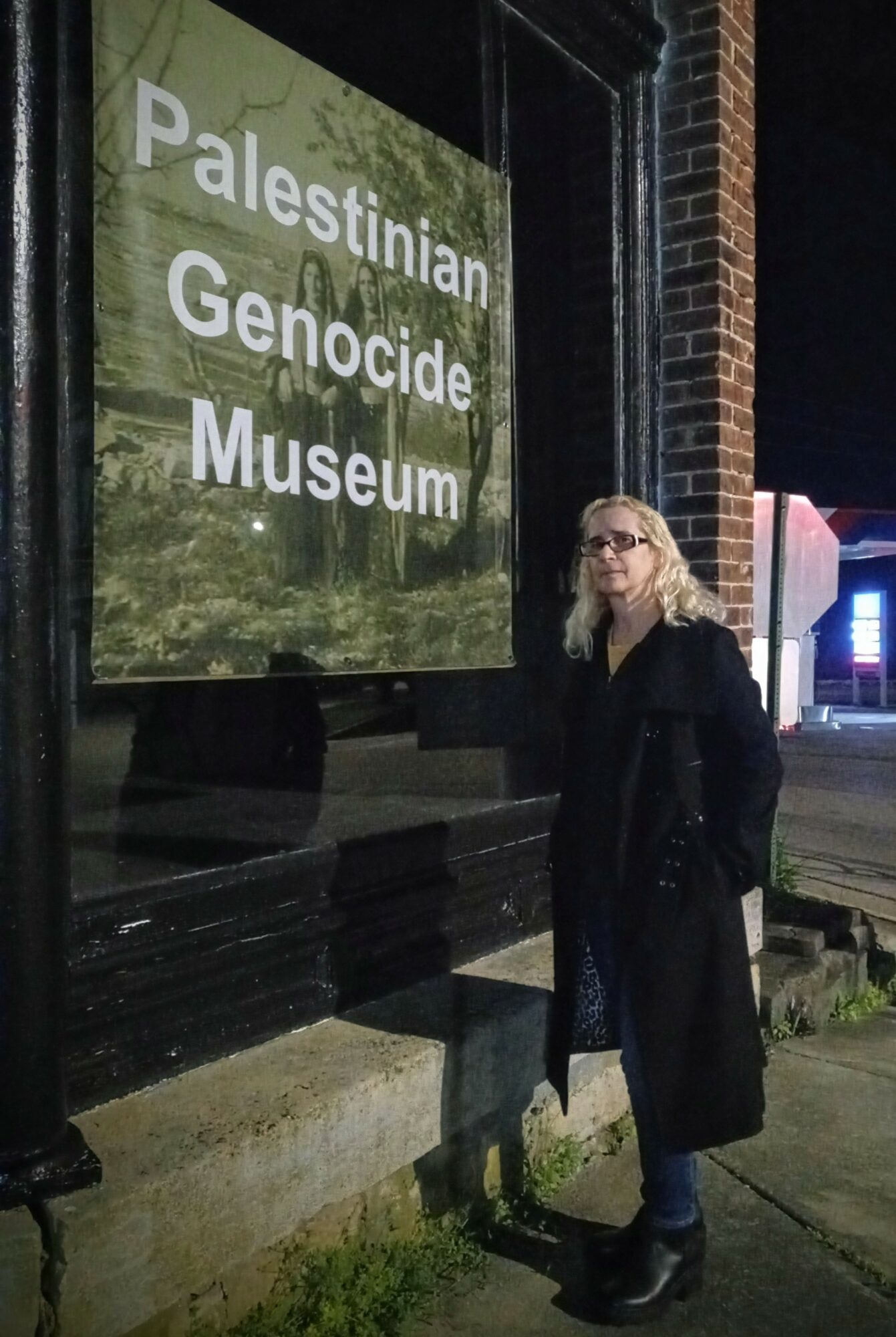 Woman standing outside Palestinian Genocide Museum at night, wearing glasses and a long coat.