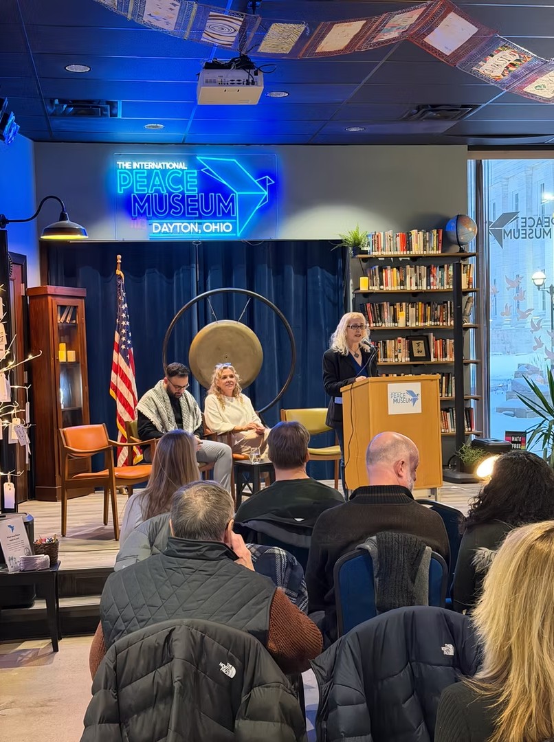 People seated in a room with a woman speaking at a podium, bookshelves, and a neon sign reading 'Museum' in Dayton, Ohio.