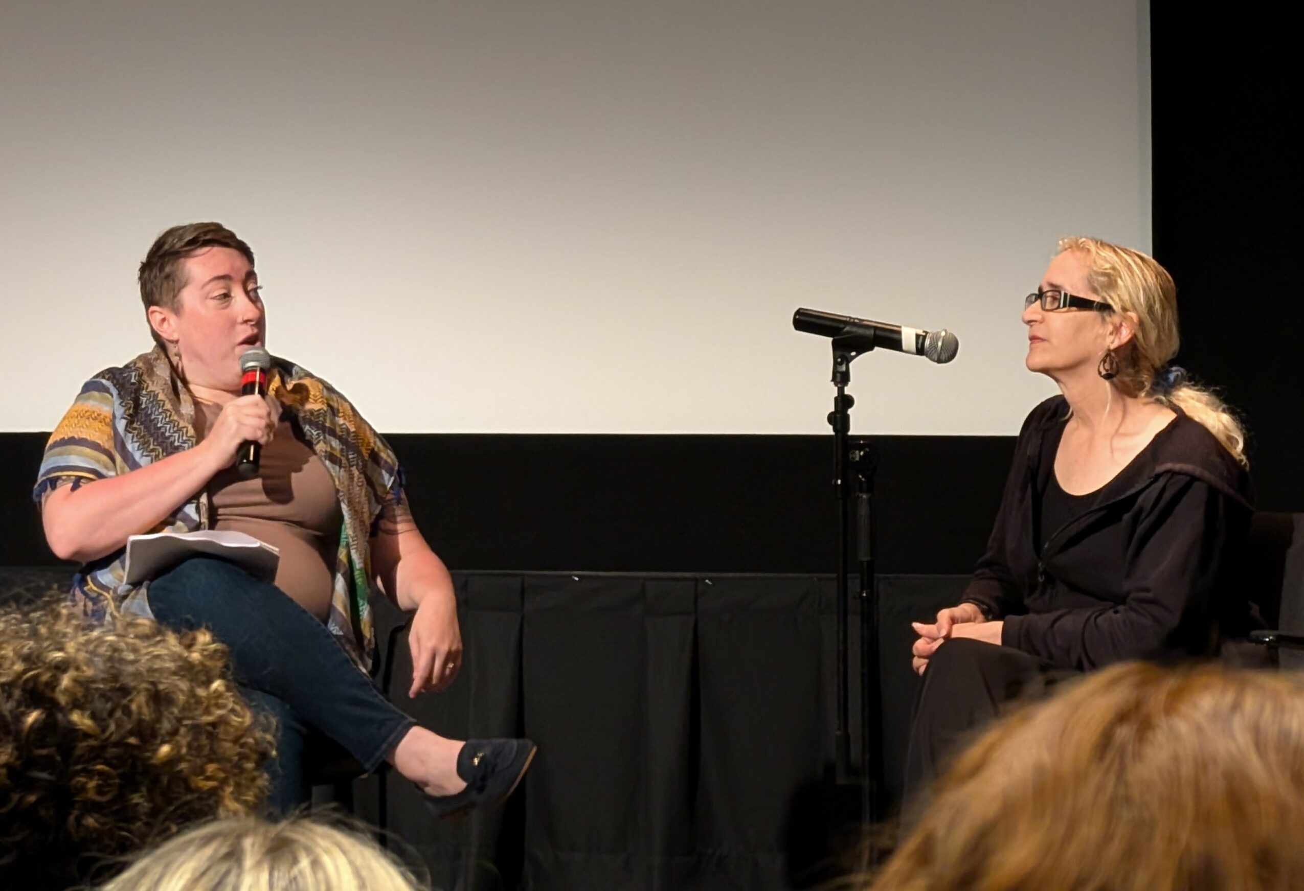 Two people sit on stage, one speaking into a microphone, the other listening, with an audience in front.