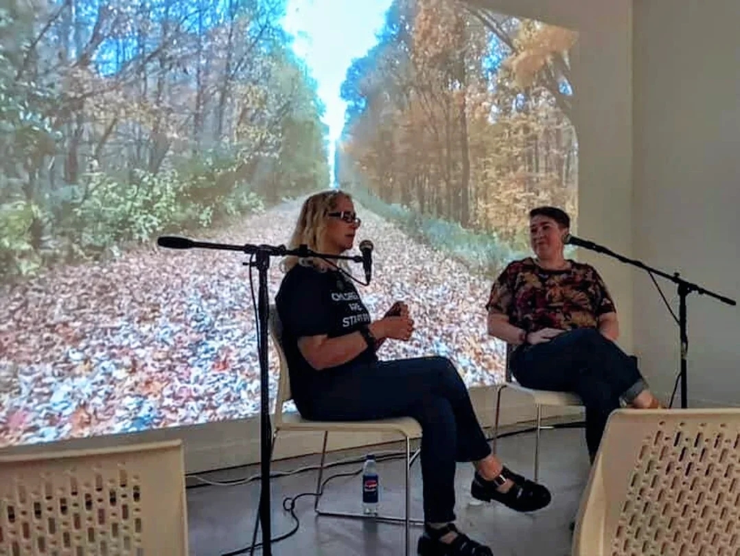 Two women sit on chairs with microphones, engaging in conversation, with a forest path background projected behind them.