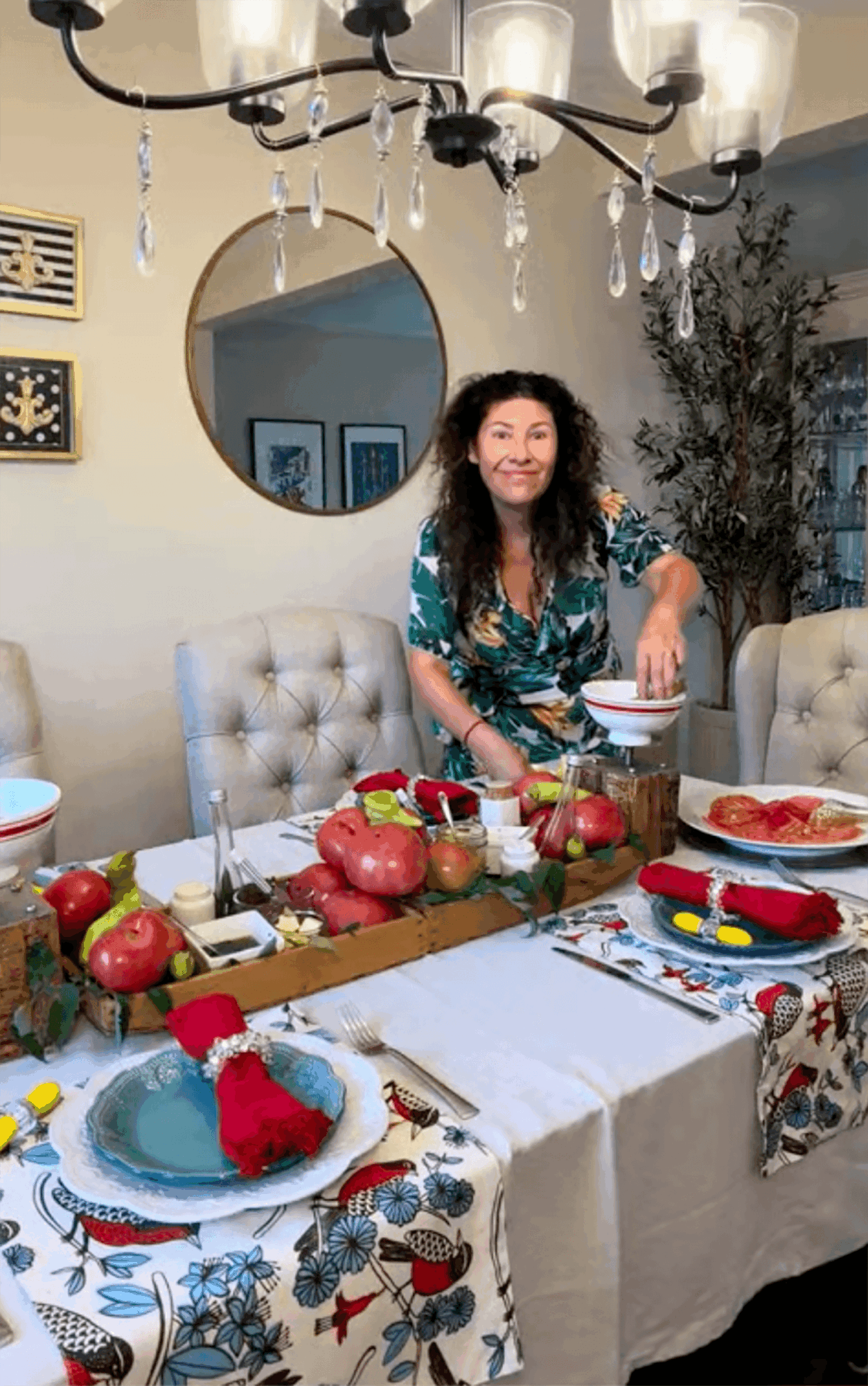 Woman with curly hair smiling at a dining table decorated with apples and festive items, in a well-lit room with a mirror and chandelier.