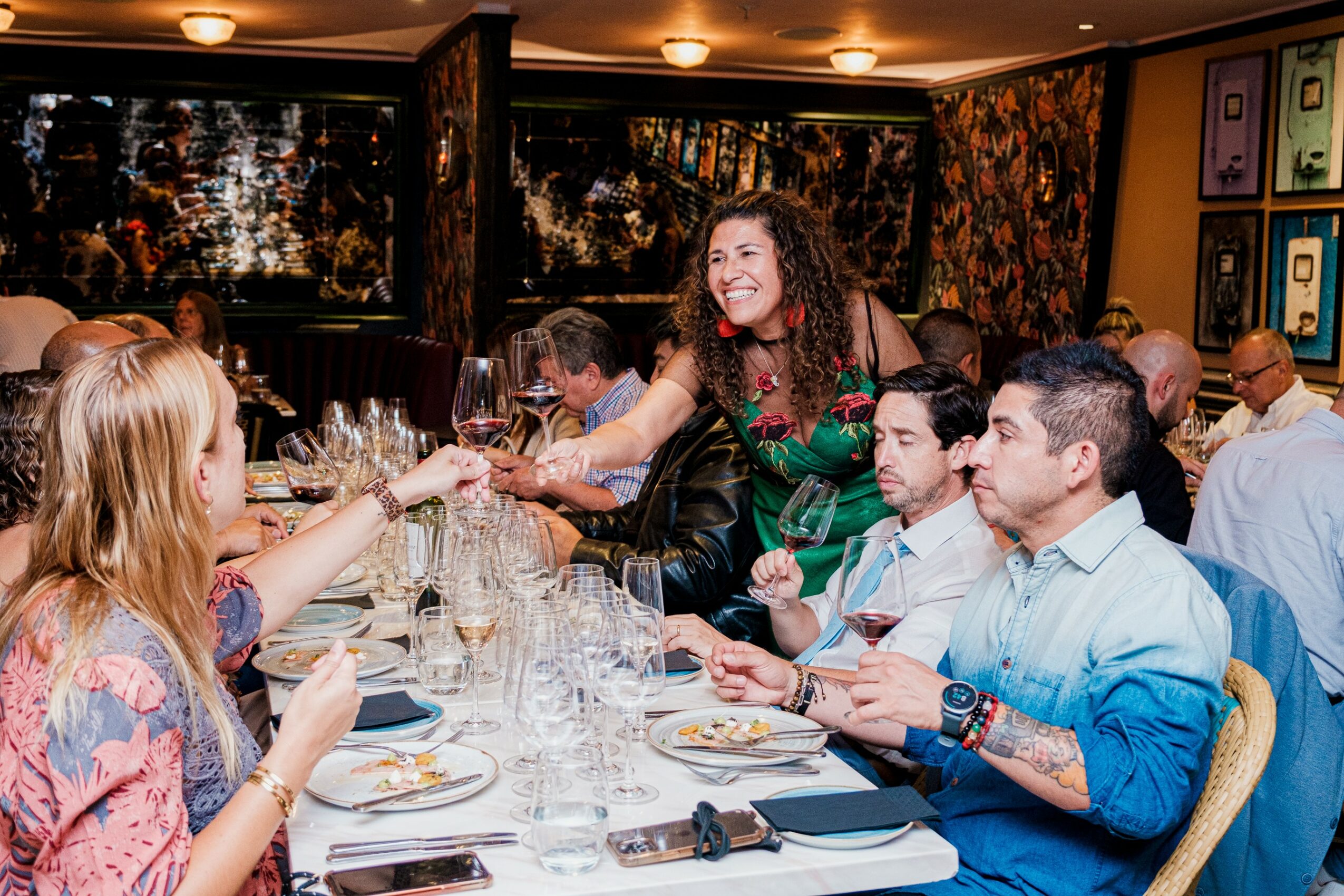 Group of people at a restaurant table, woman handing a glass to another woman, smiling, with others dining around.
