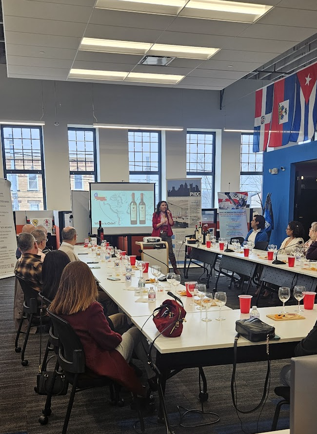 People seated around a conference table in a room with large windows, flags, and a presentation screen, listening to a speaker.