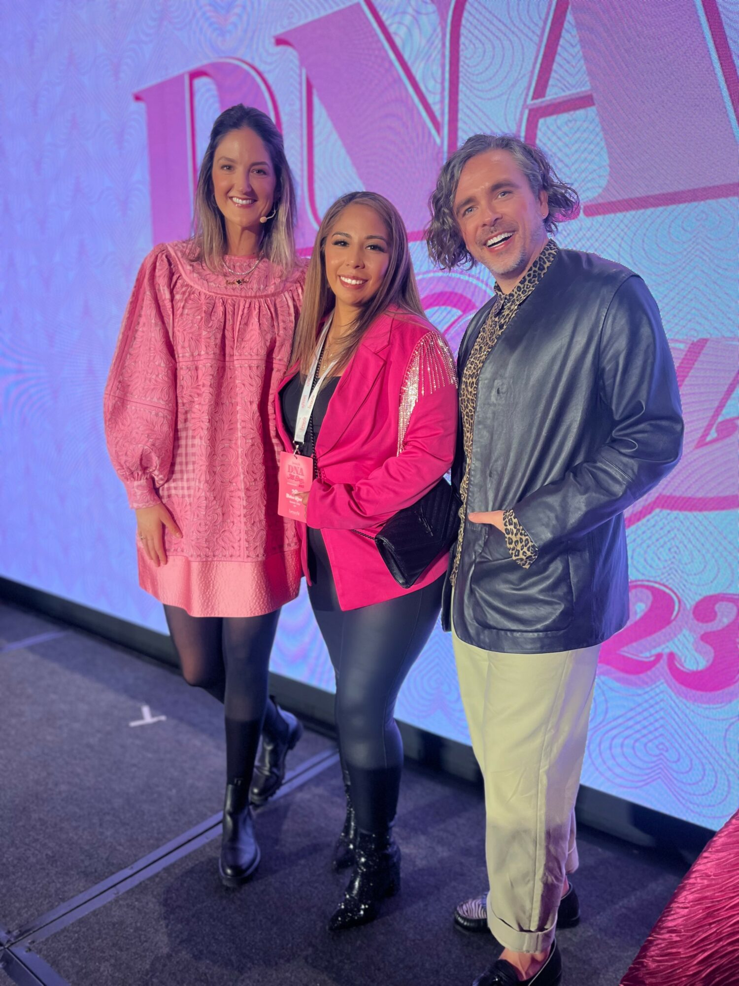 Three people standing together in front of a colorful background, smiling, with a large pink and purple sign behind them.