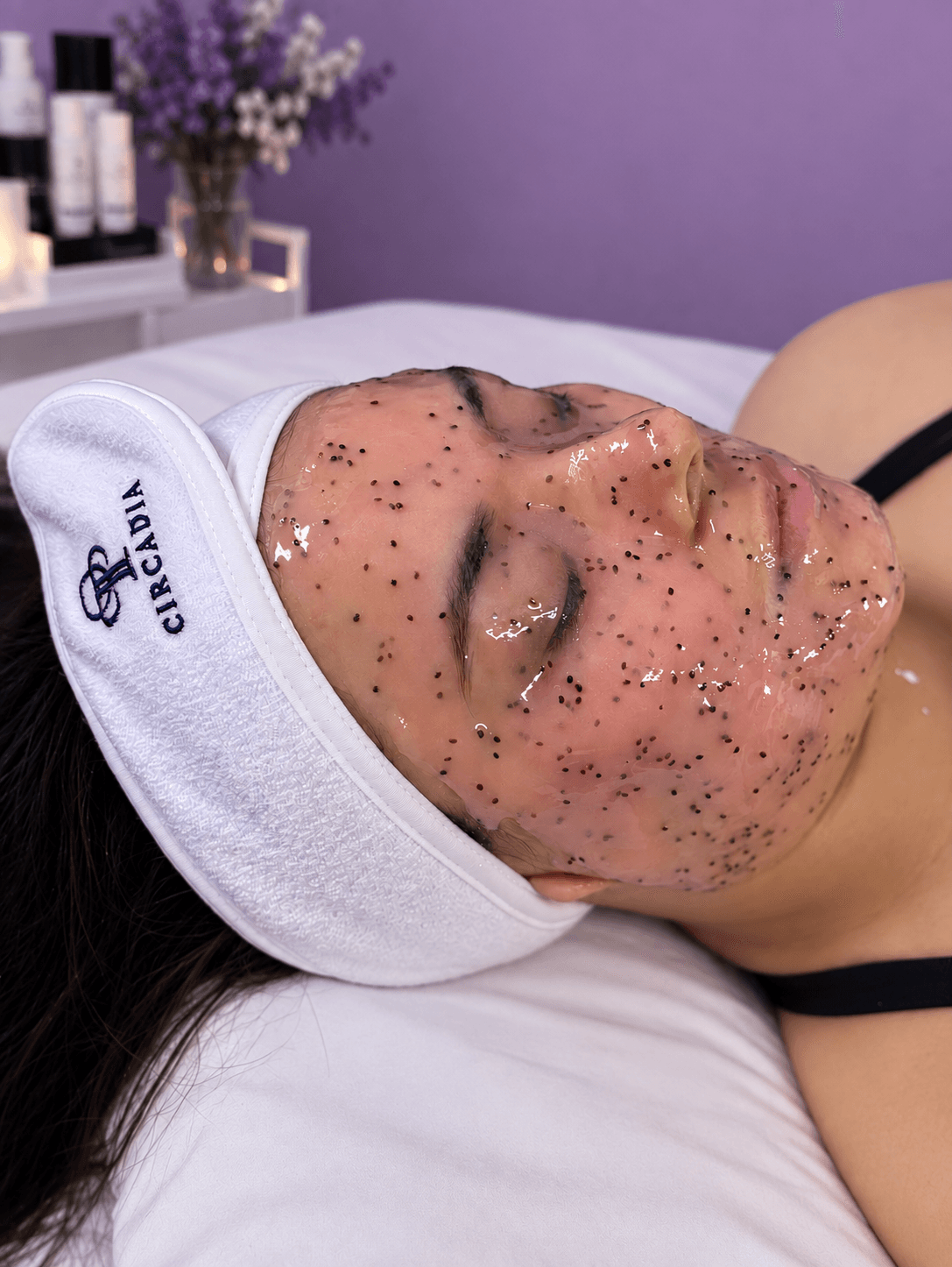Person lying on bed with face covered in glitter, wearing a white headband and black top, in a room with purple wall and flowers.