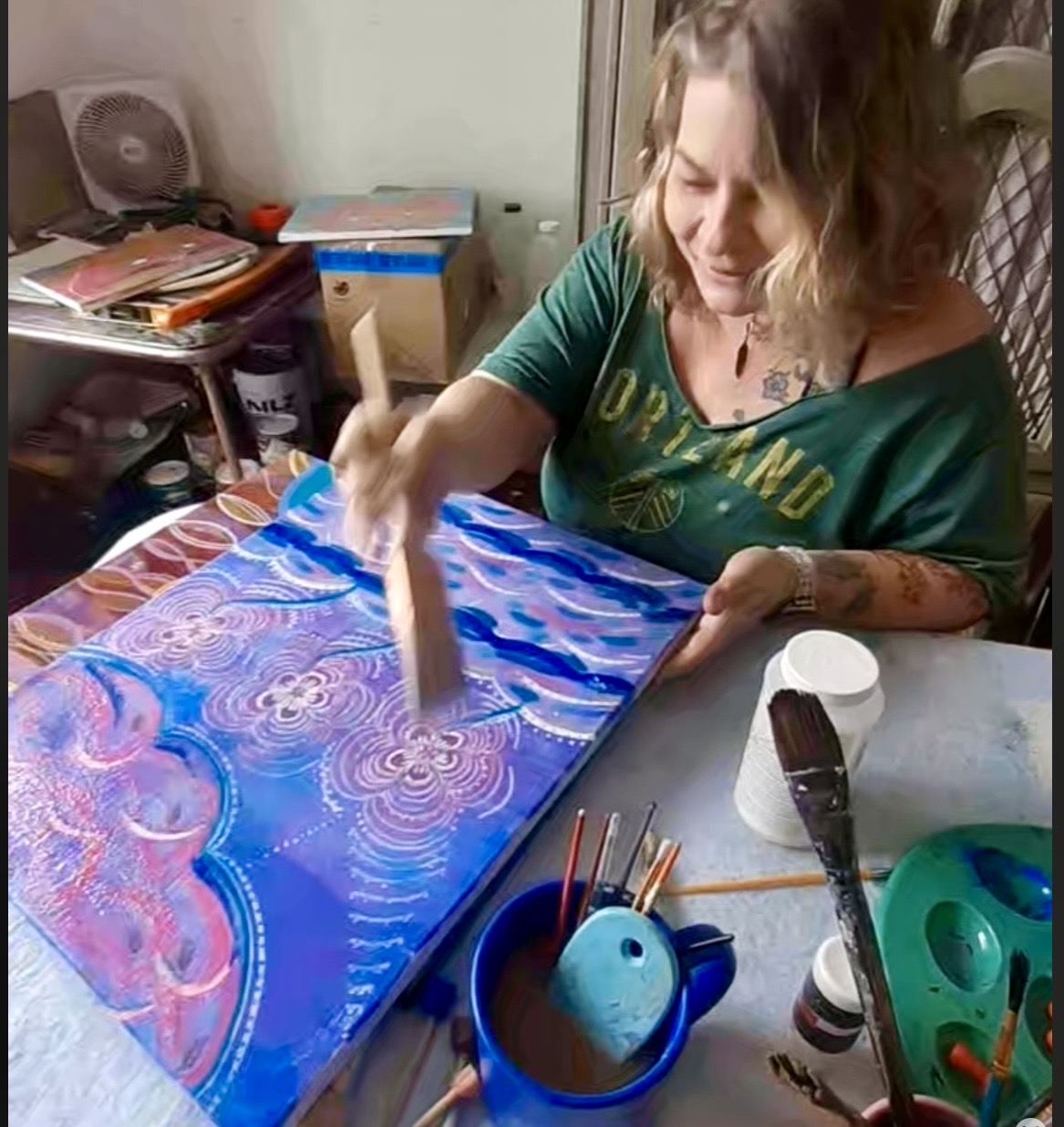 Woman painting a colorful abstract design on a canvas at a table, with art supplies nearby.