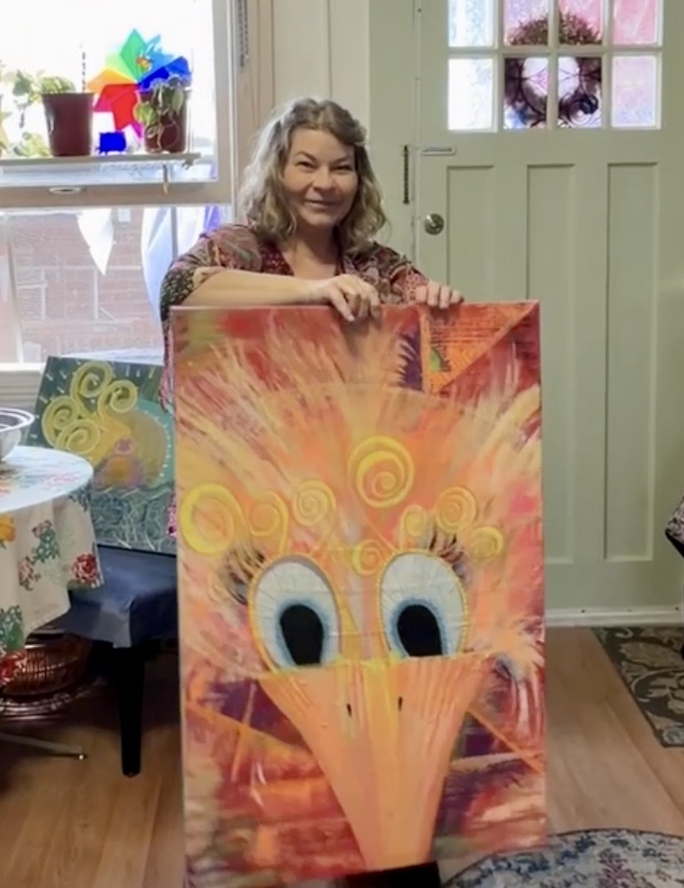Woman holding a colorful painting of a cartoonish bird with large eyes and a beak, indoors near a door and window.