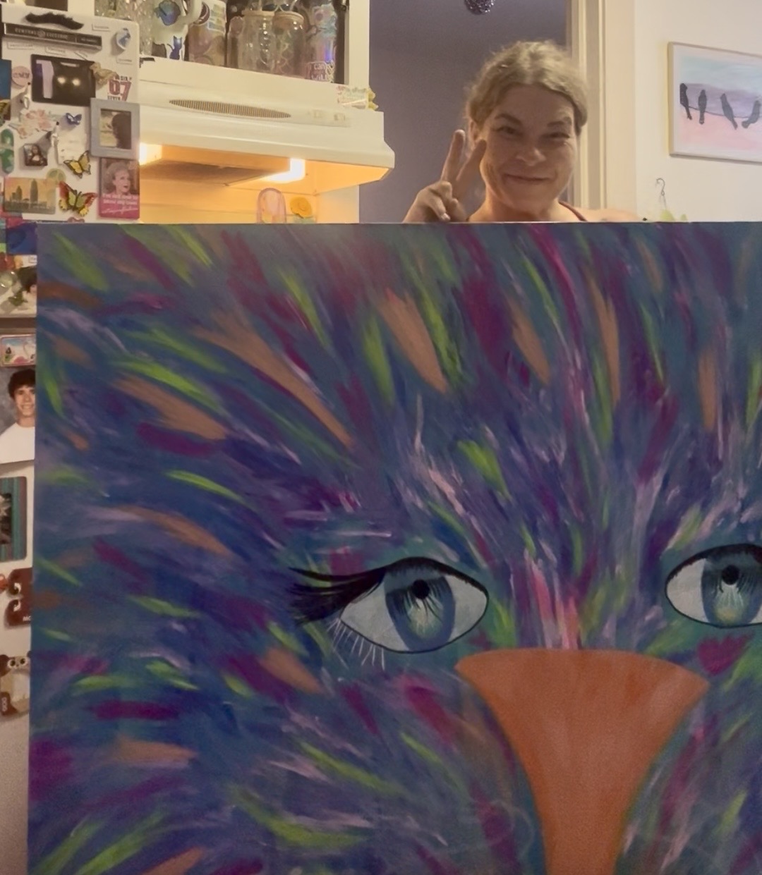 Person smiling and making a peace sign behind a colorful abstract painting of a cat's face with blue eyes and an orange nose.