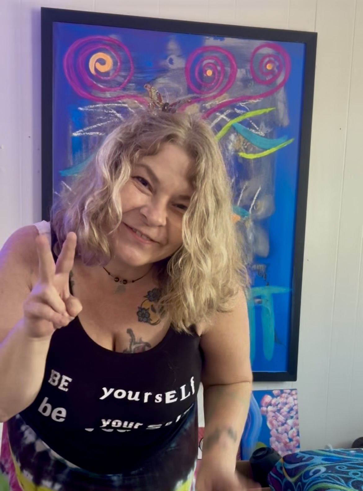 Smiling woman with curly blonde hair making a peace sign in front of colorful abstract artwork.