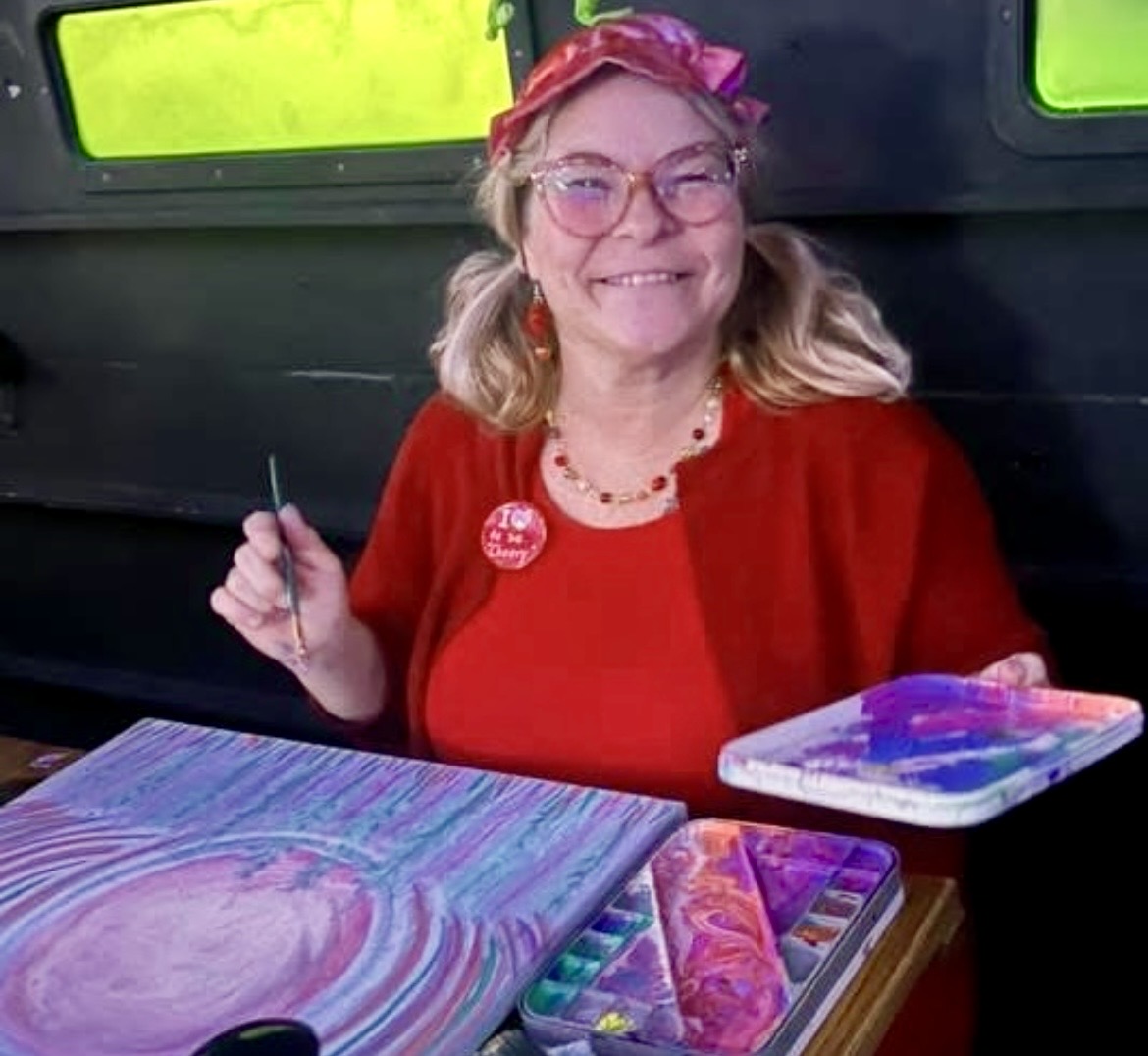 Smiling woman with glasses, wearing a red top and pink headband, sitting at a table with colorful art supplies.