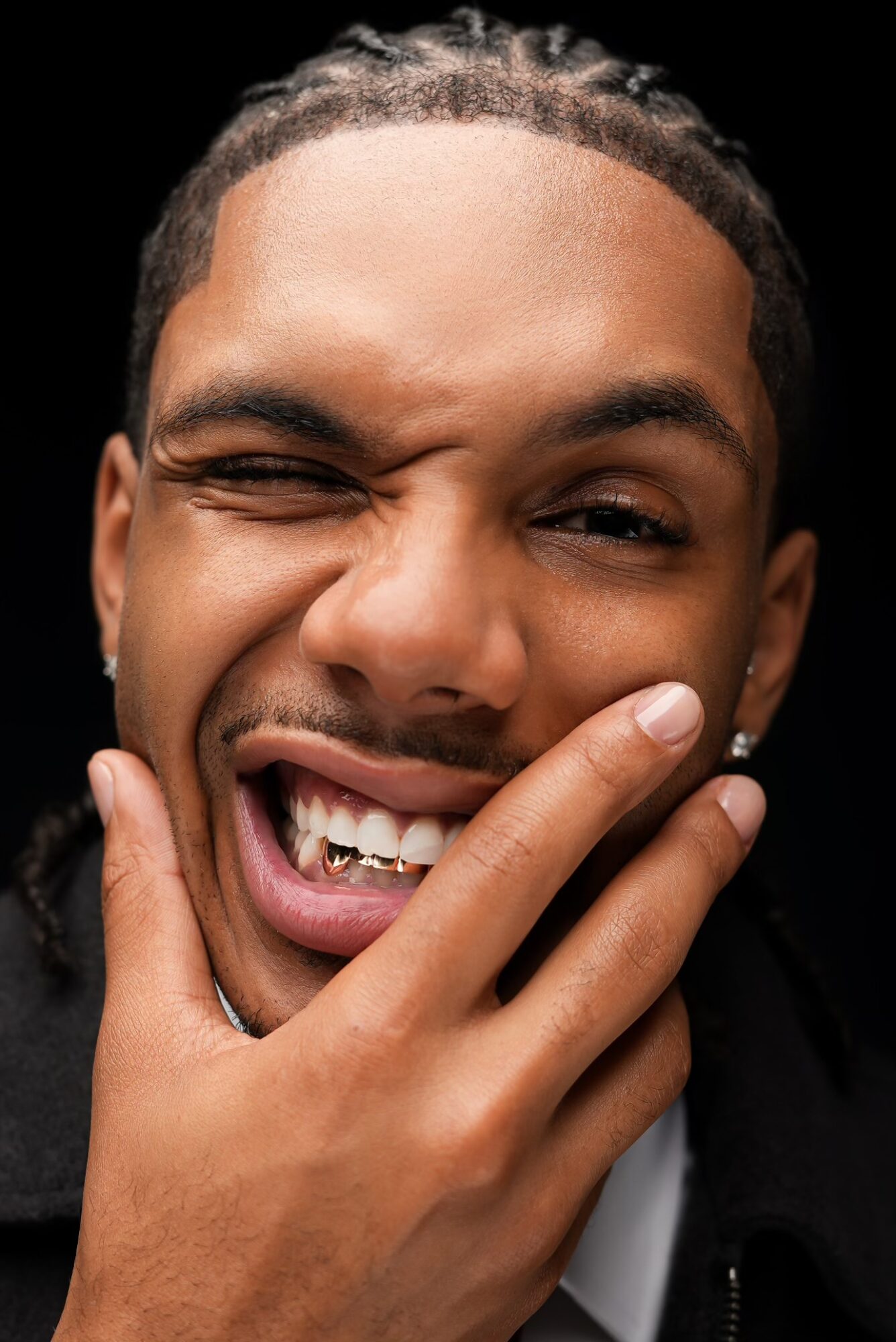 Person with braided hair winking and smiling, hand touching face, black background