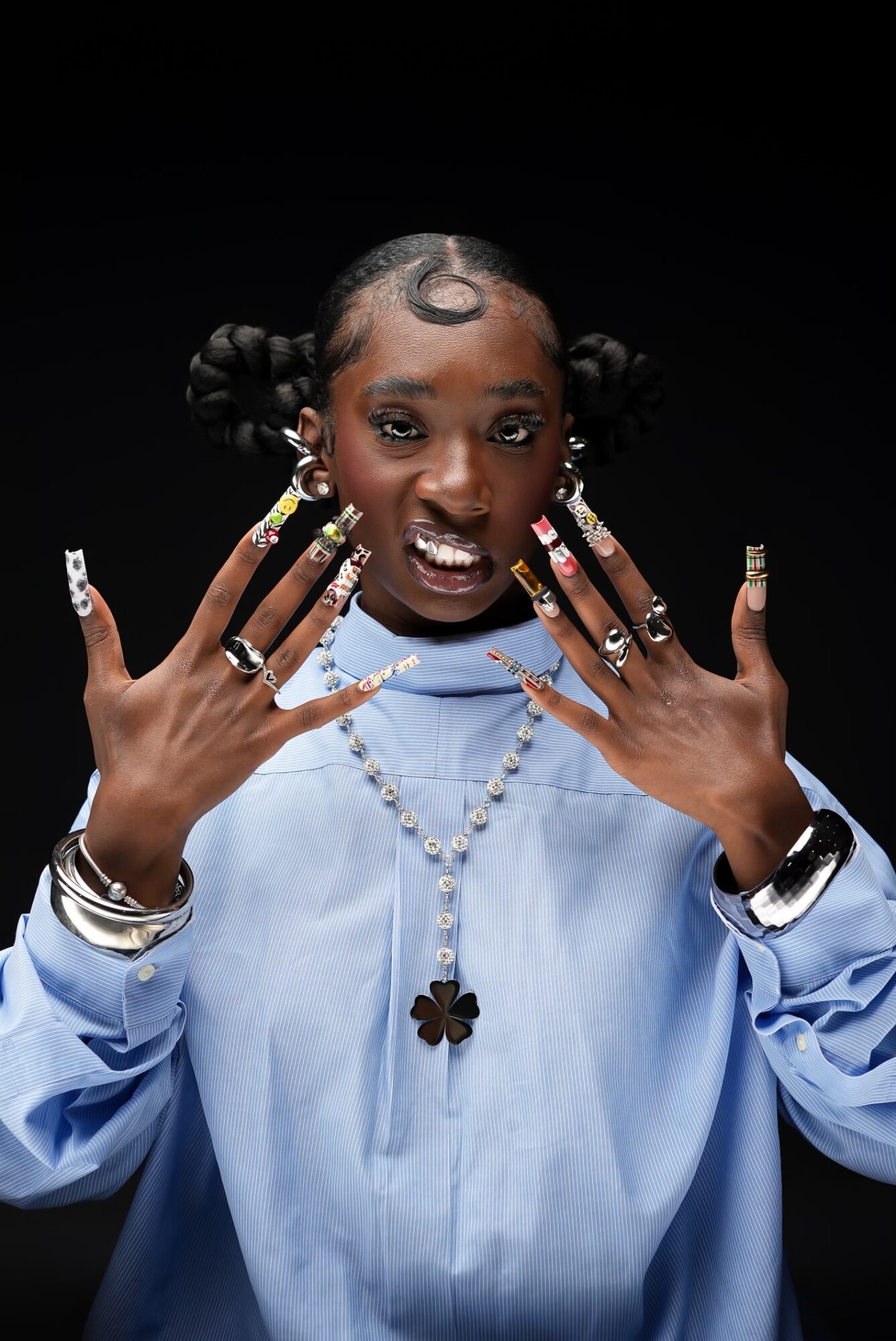 Young person with styled hair, jewelry, and a blue shirt, posing with hands near face against a black background.