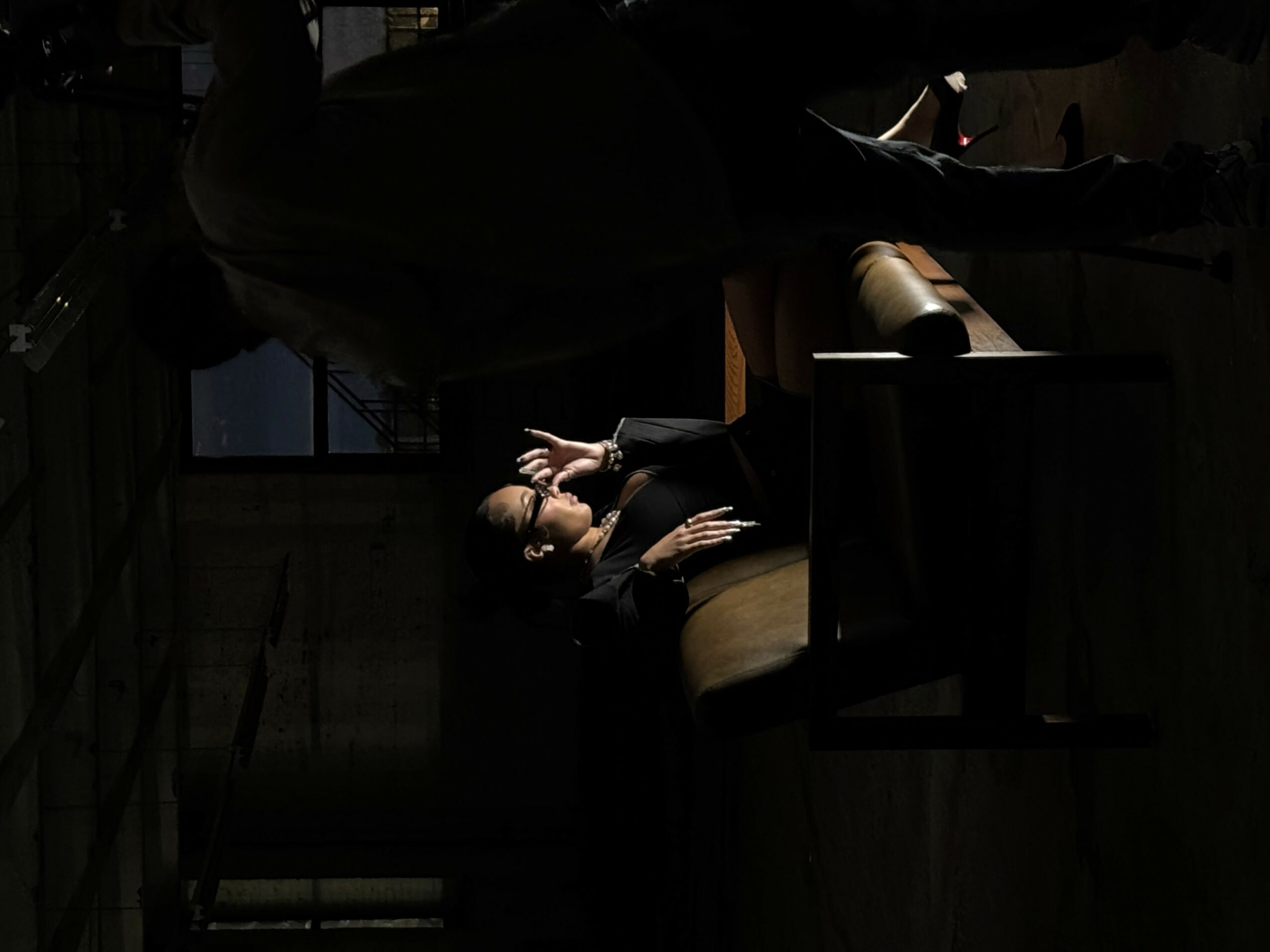 Person sitting at a table in a dimly lit room, with a window in the background, head resting on hand.