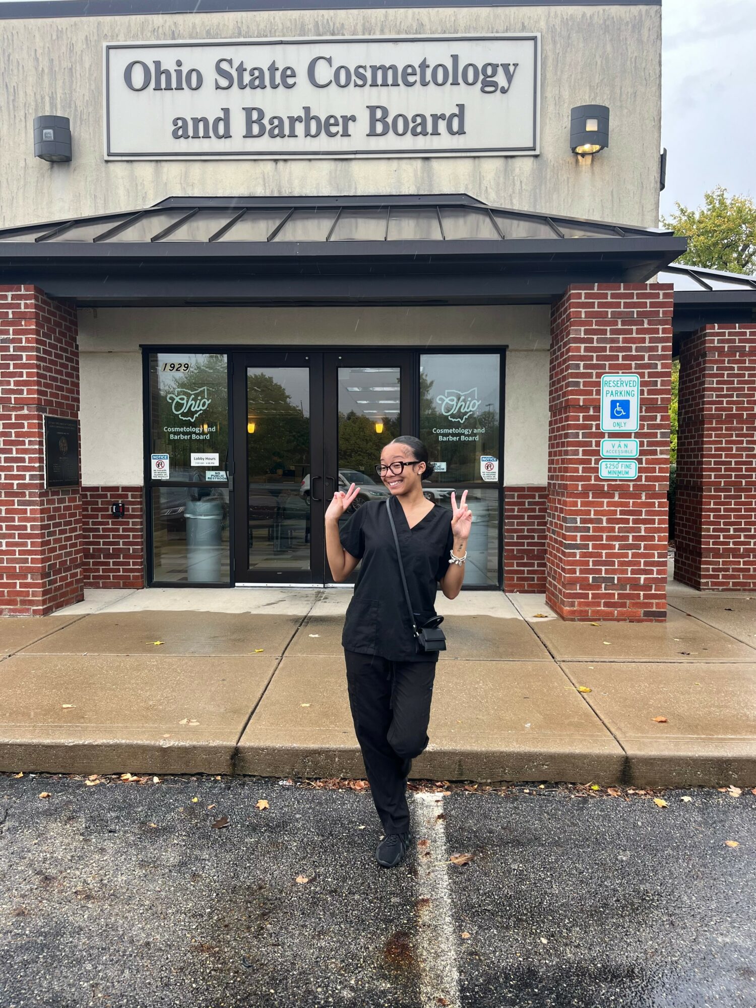 Woman standing outside Ohio State Cosmetology and Barber Board building, smiling, holding a phone, on a rainy day.