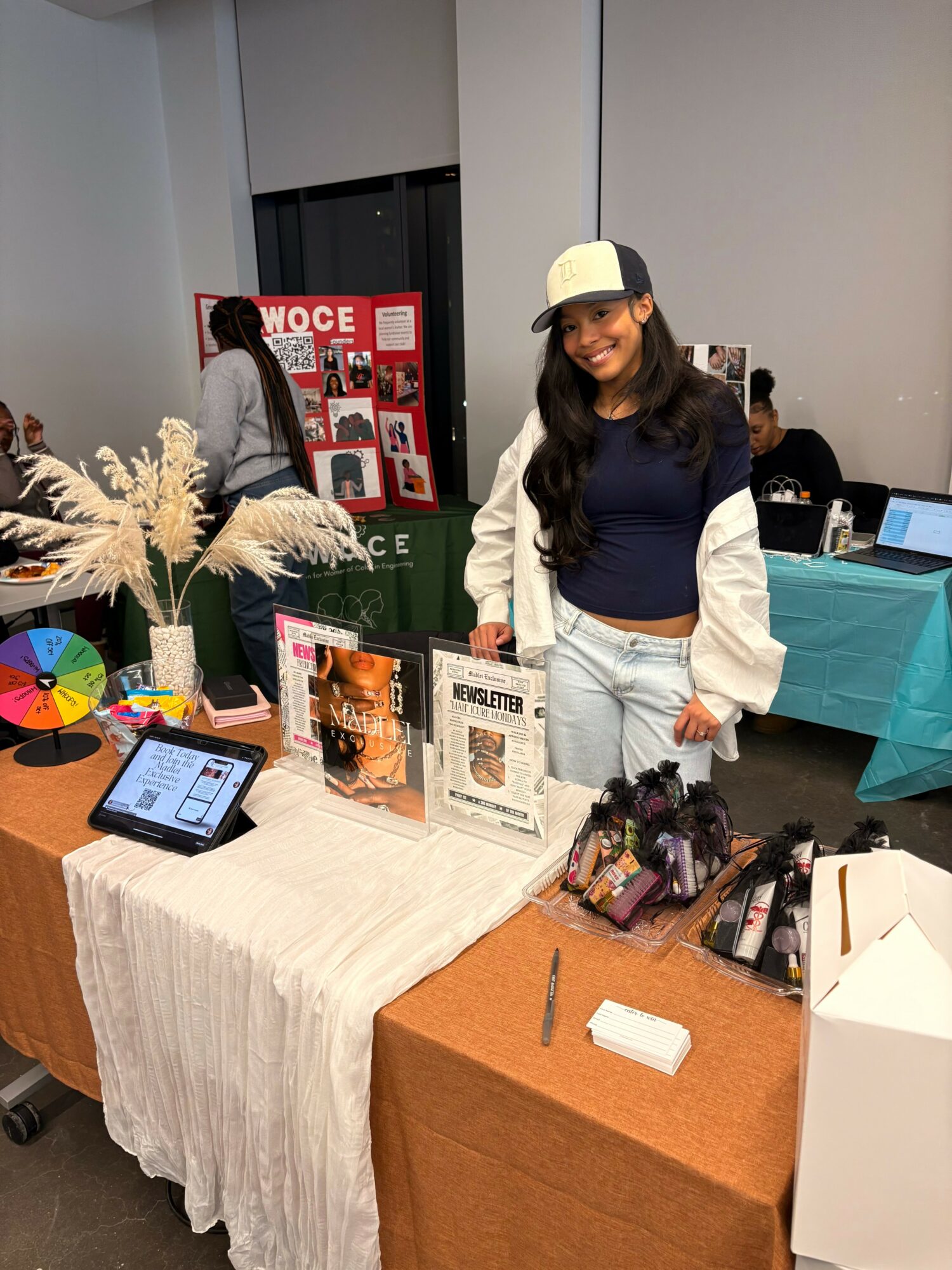 Young woman with long dark hair, wearing a cap, navy sweater, and light jeans, stands behind a table at an indoor event.