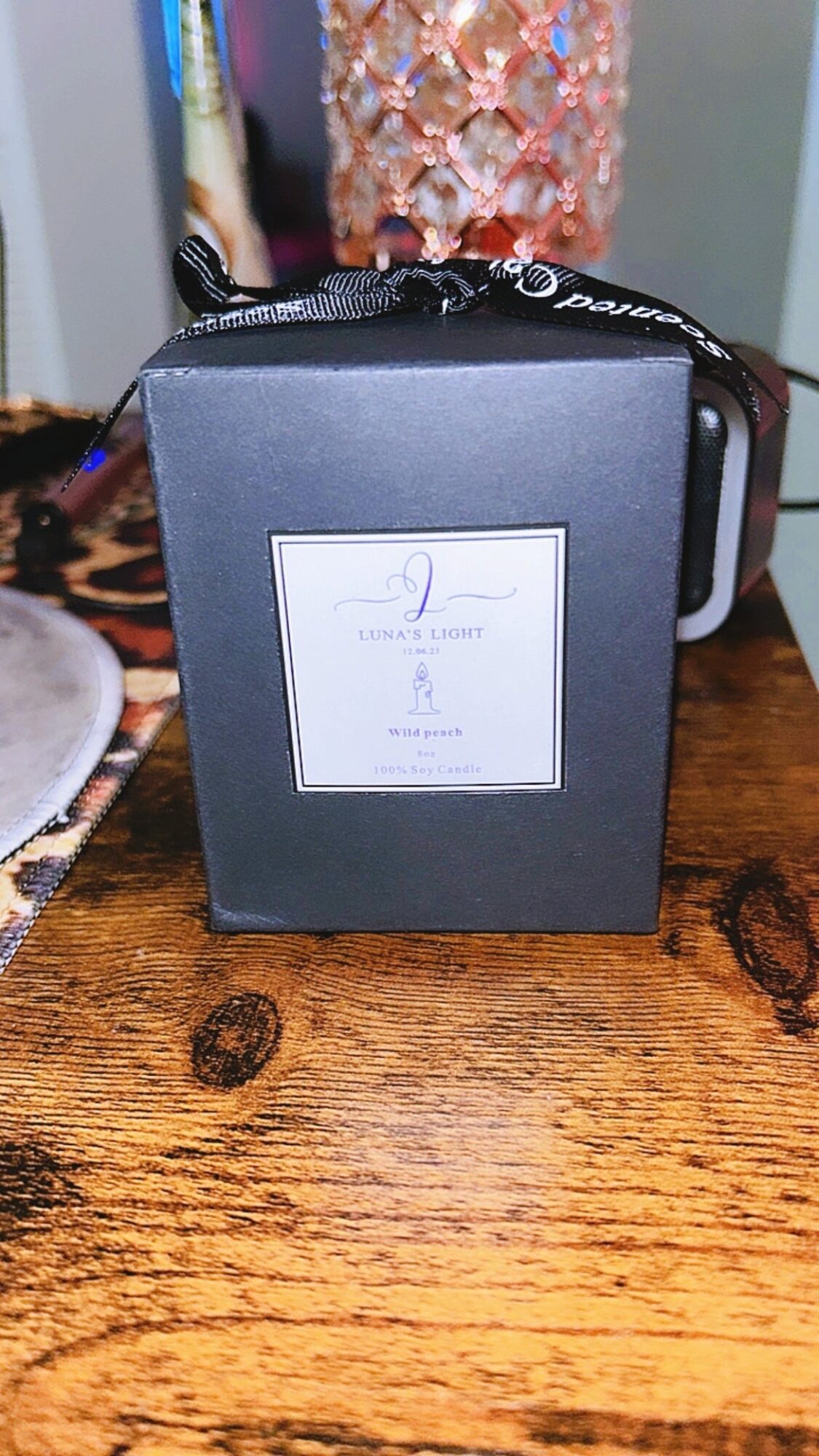 Black box with a white label on a wooden surface, surrounded by various objects, including a pink decorative item and a ribbon.