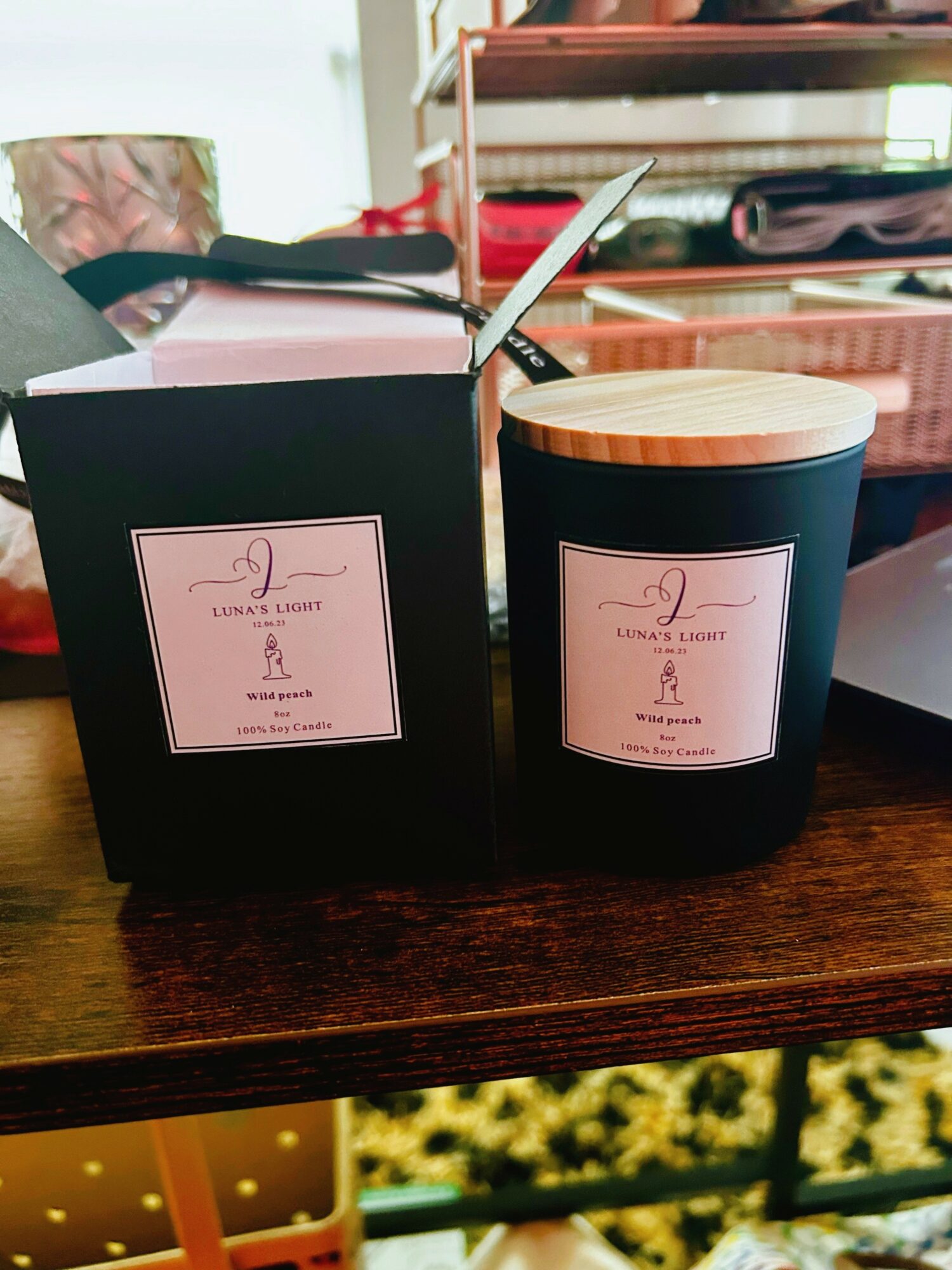 Black candle jar with a wooden lid and a pink label, next to a black box with a similar label, on a wooden surface.