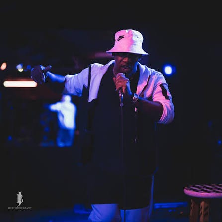 Person wearing a white bucket hat and dark jacket singing into a microphone on stage, with a blurred background and blue lighting.