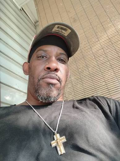Man wearing a cap and a cross necklace, looking at the camera, with a background of a wall and ceiling.