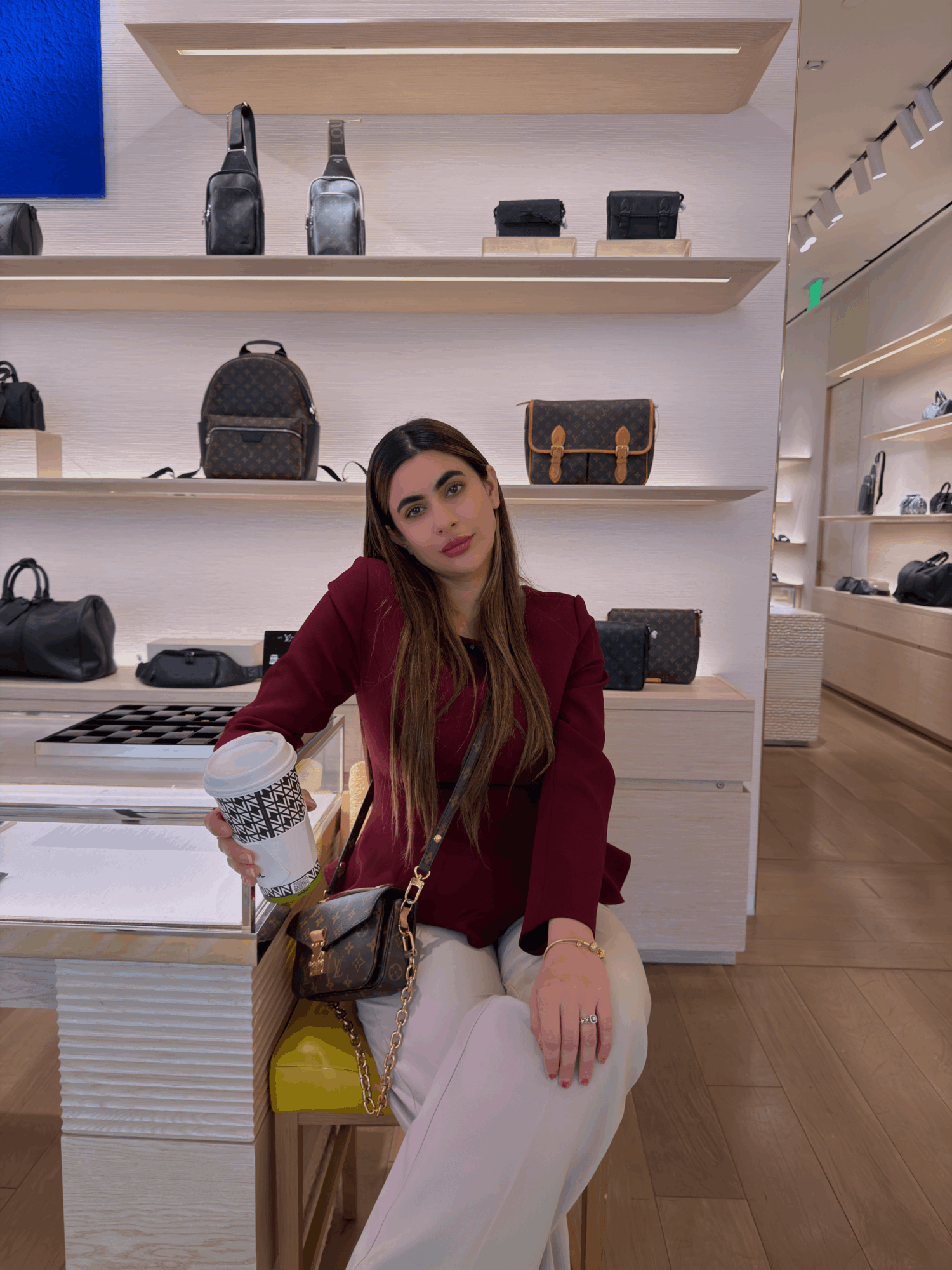 Young woman with long hair sitting in a store, surrounded by shelves of handbags and bottles, holding a coffee cup.