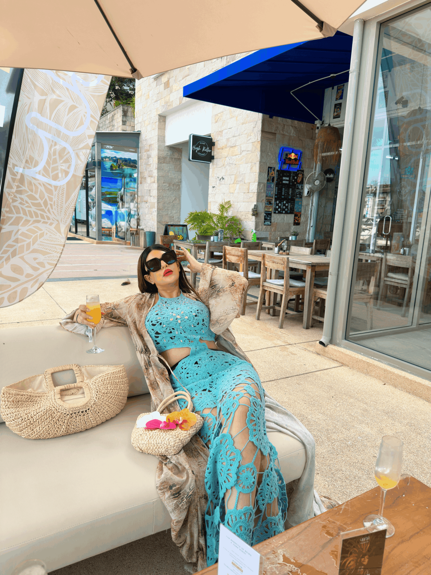 Woman in sunglasses and a blue dress relaxing outdoors under umbrellas, with a table and chairs nearby.
